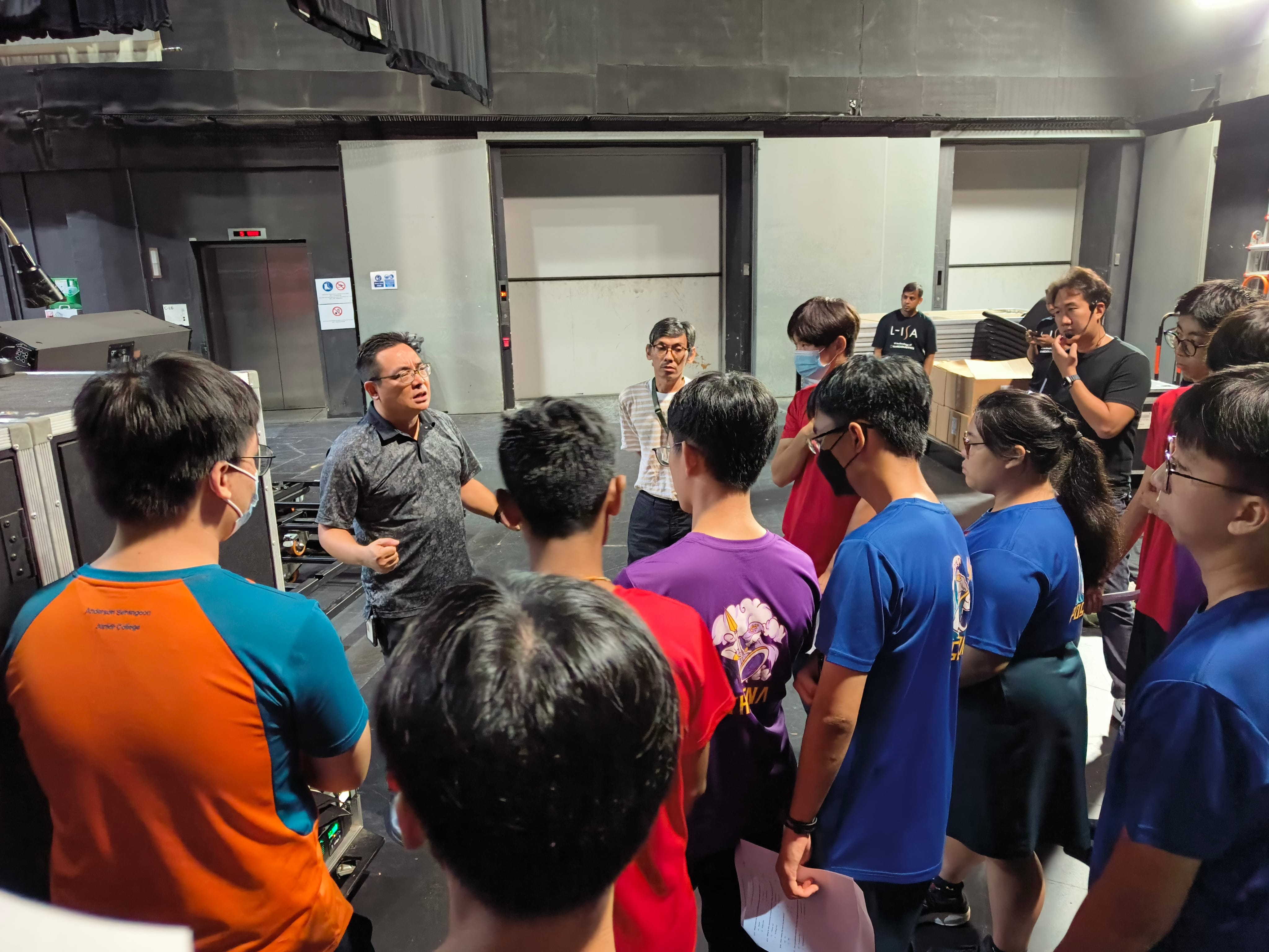 A man addresses a group of young people gathered in a black-walled indoor studio or theater space.