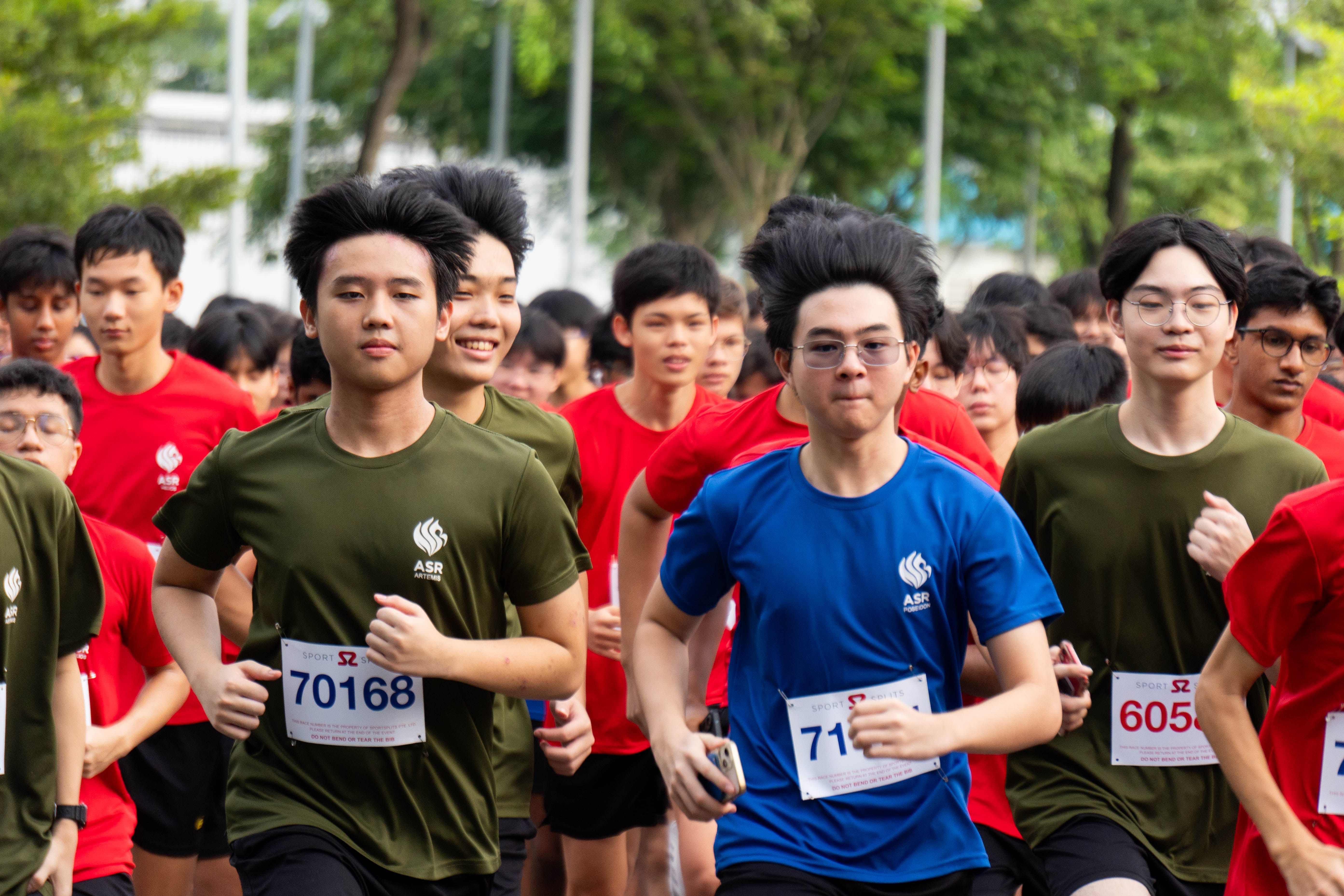 A large group of young runners wearing red, green, and blue ASR shirts with race bibs competing outdoors.
