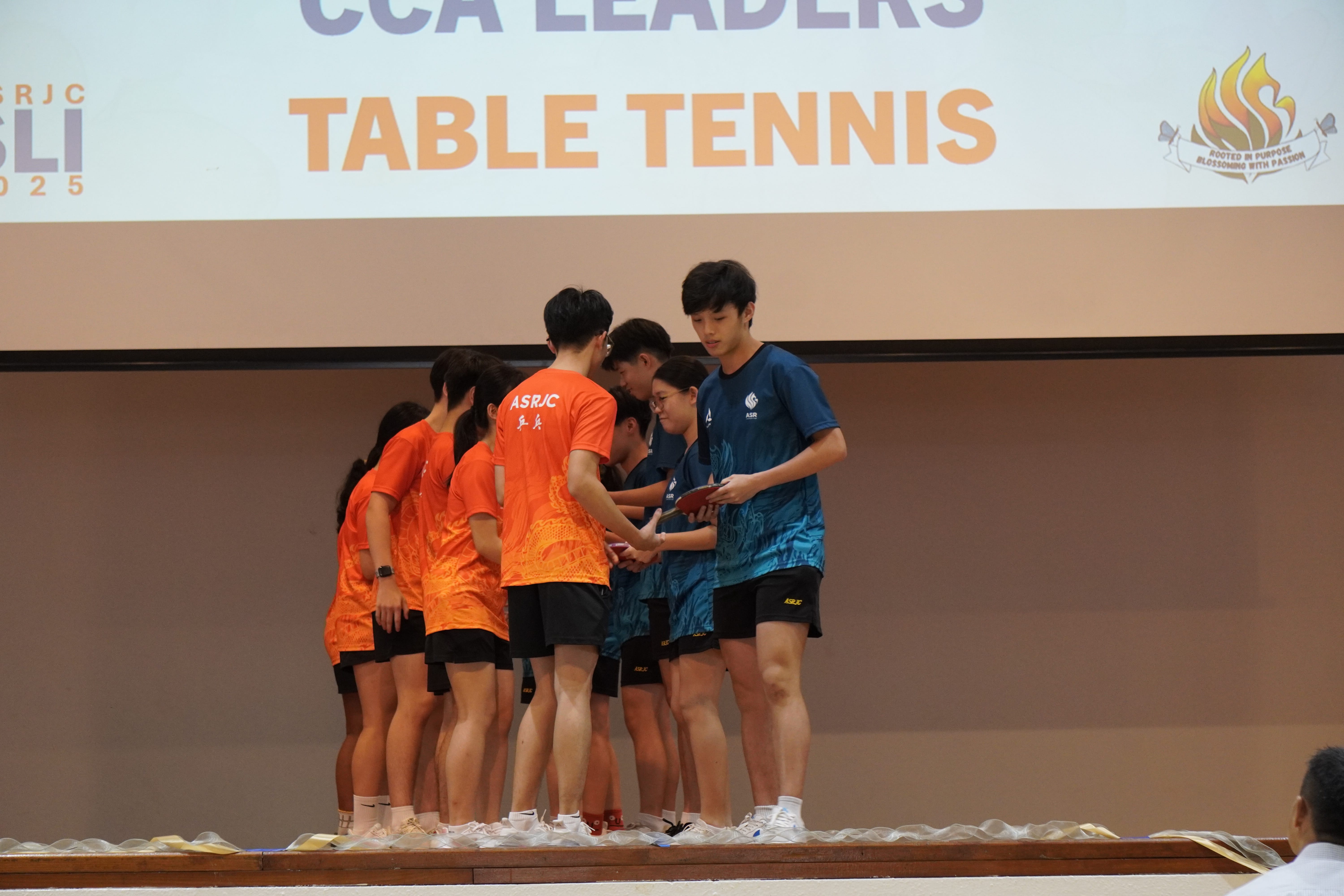 Two teams in orange and blue table tennis jerseys huddle onstage under a banner reading TABLE TENNIS.