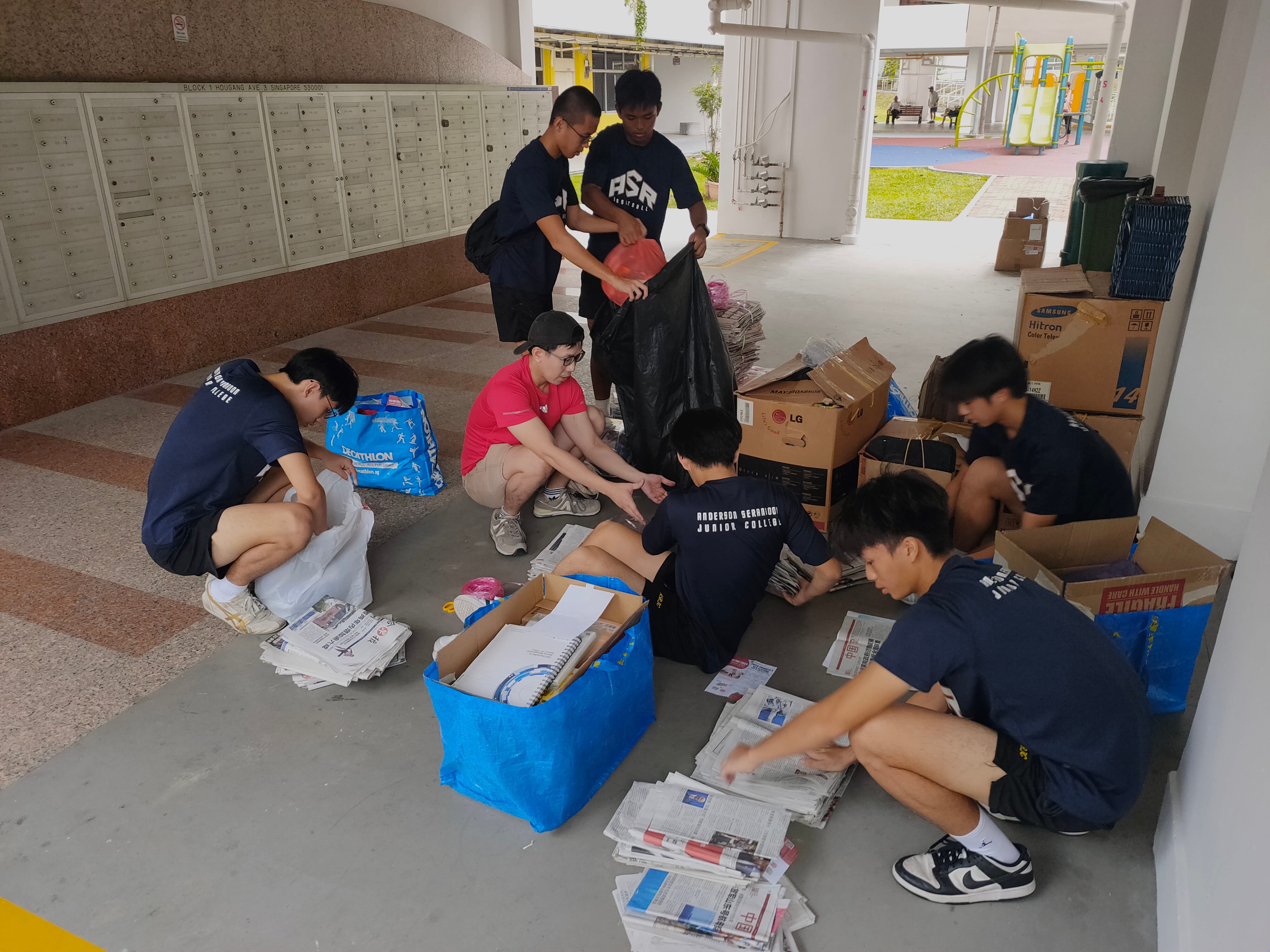 Several teenagers sort newspapers and cardboard boxes on the ground floor of a residential block near mailboxes.
