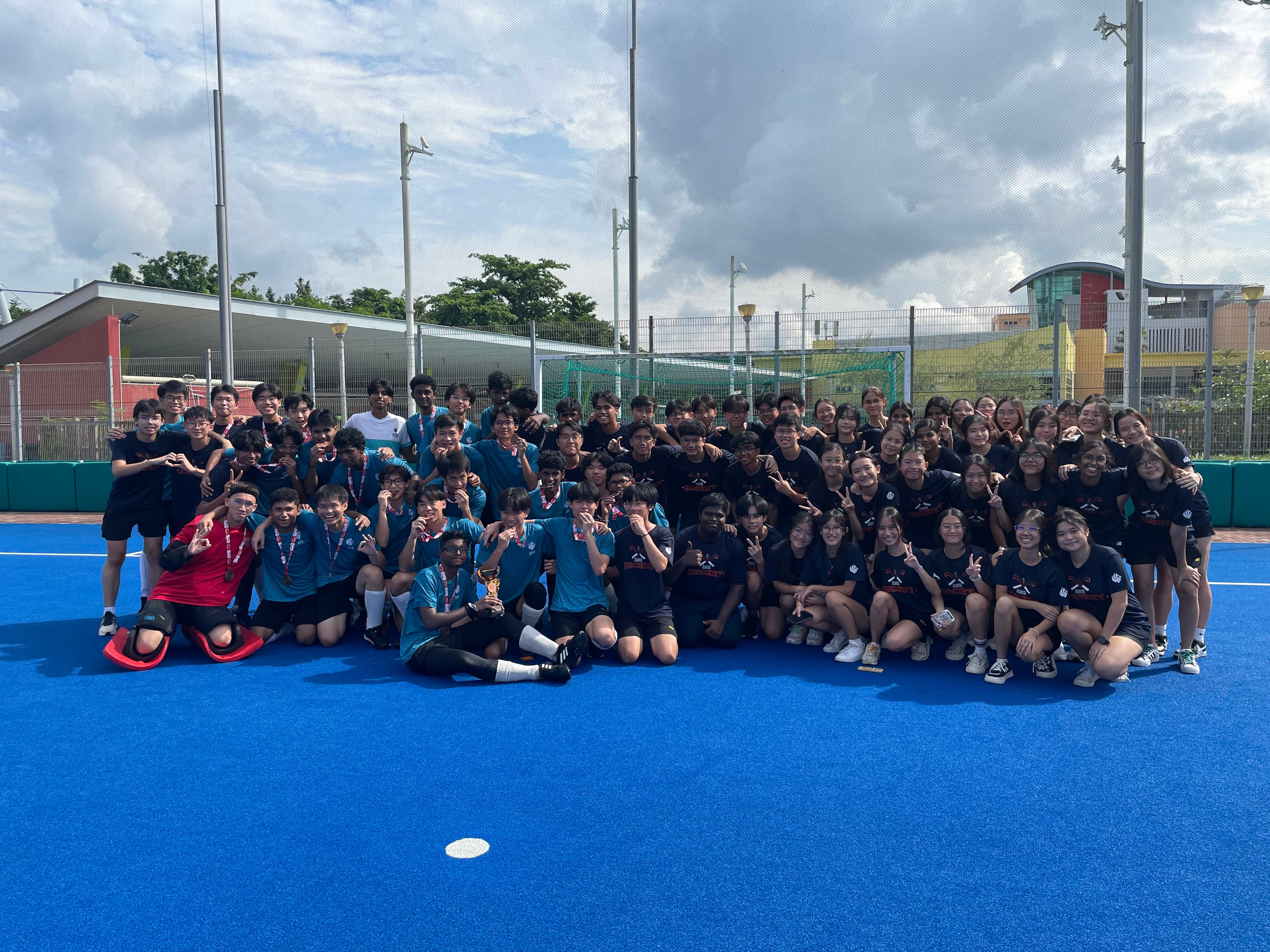 Large group of students in blue and black shirts posing on a blue sports court, some holding medals and a trophy.