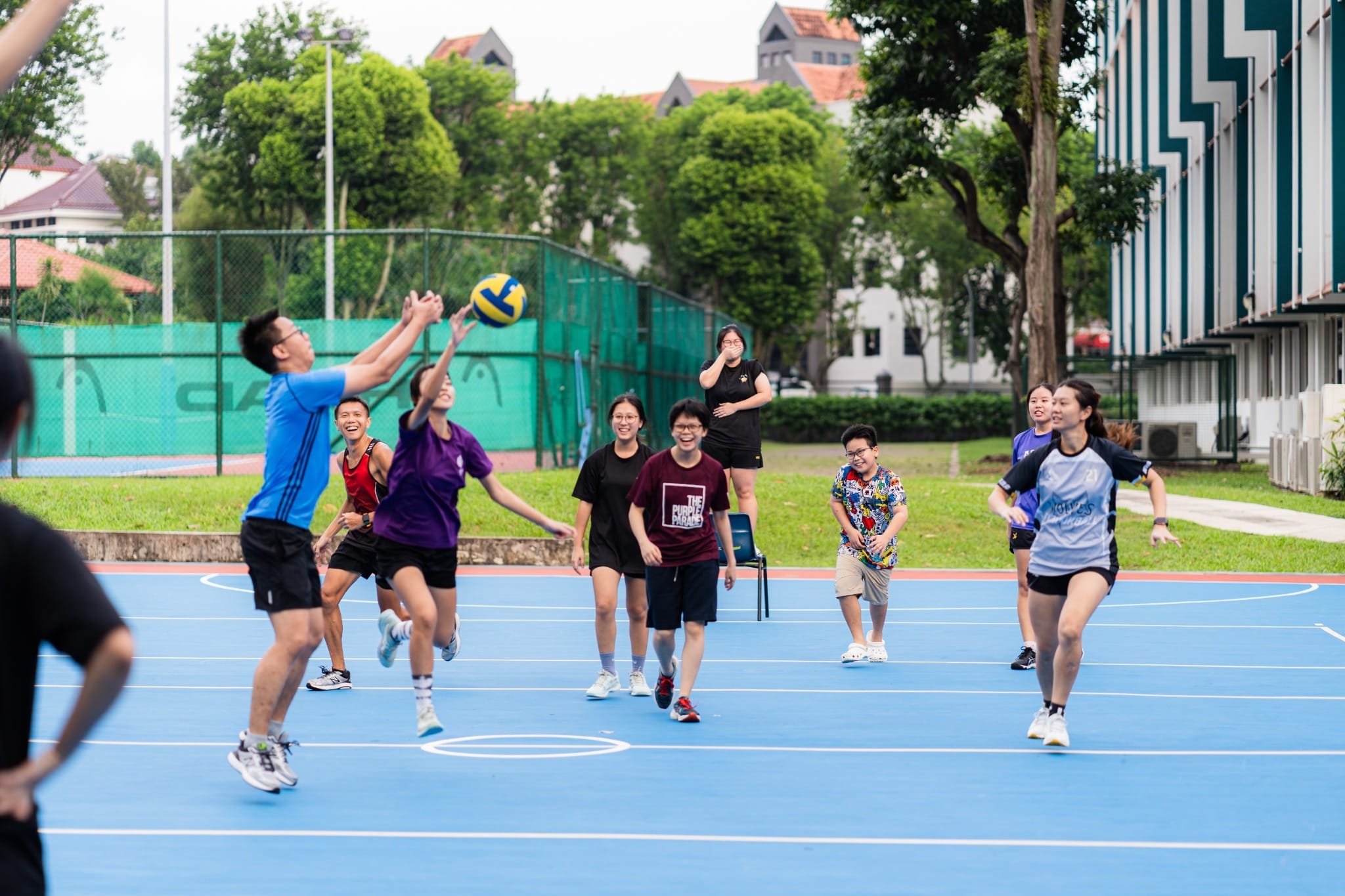 A group of young people playing volleyball on a blue outdoor court, with one player jumping to hit the ball.