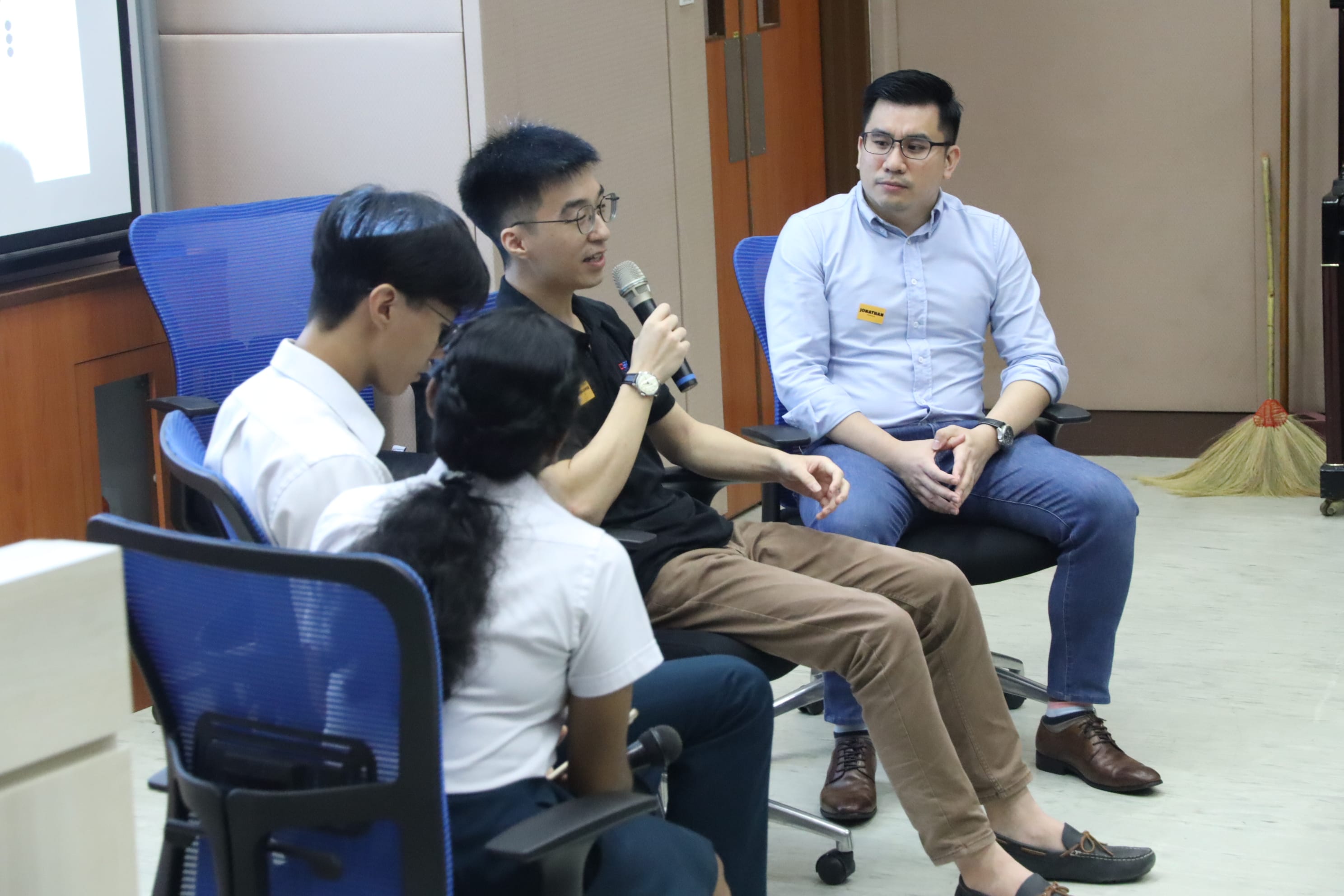 Four people sit in chairs during a panel discussion; one man speaks into a microphone while others listen.