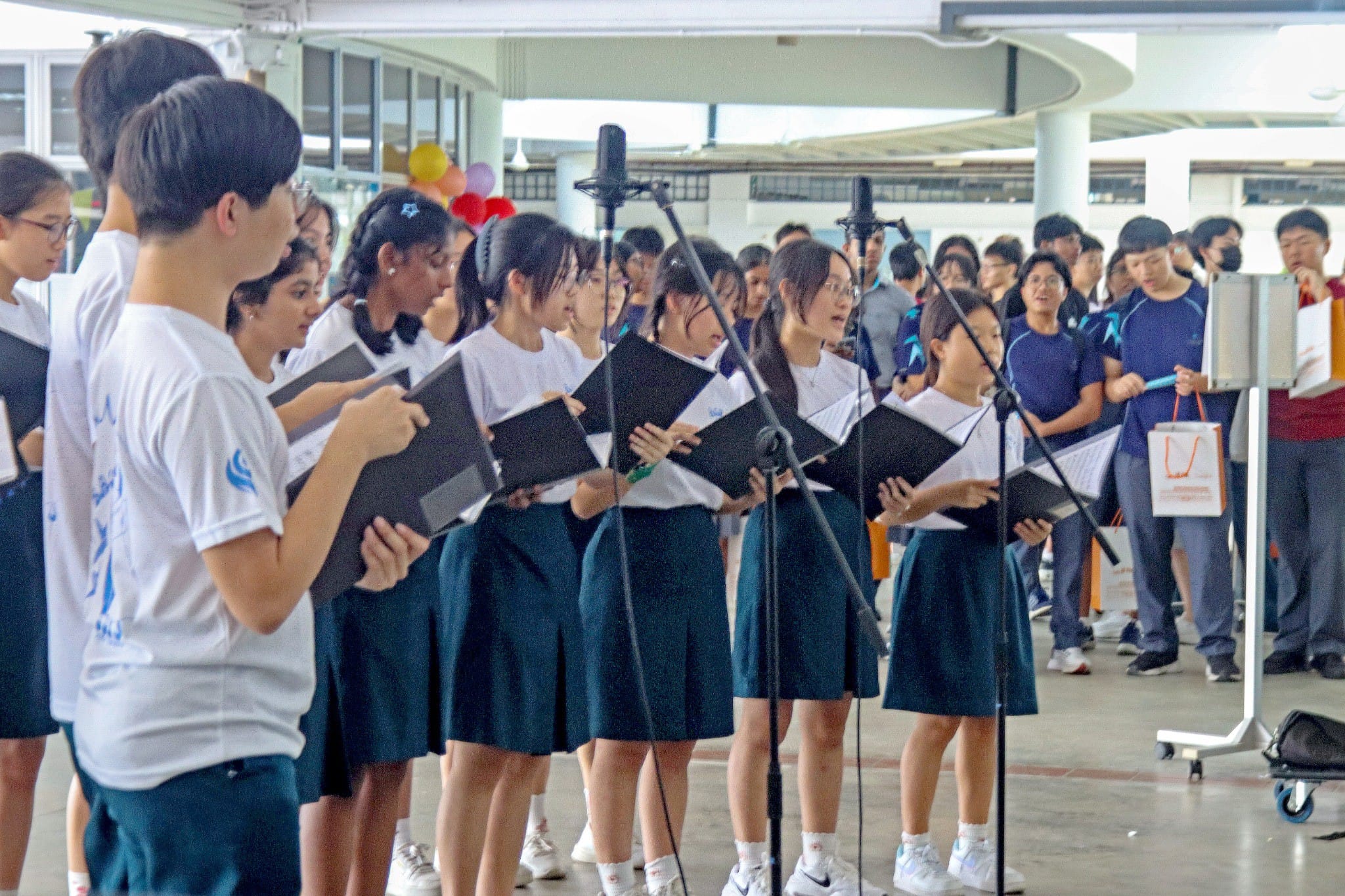 A school choir of students in white shirts and dark skirts singing from black folders near microphone stands indoors.
