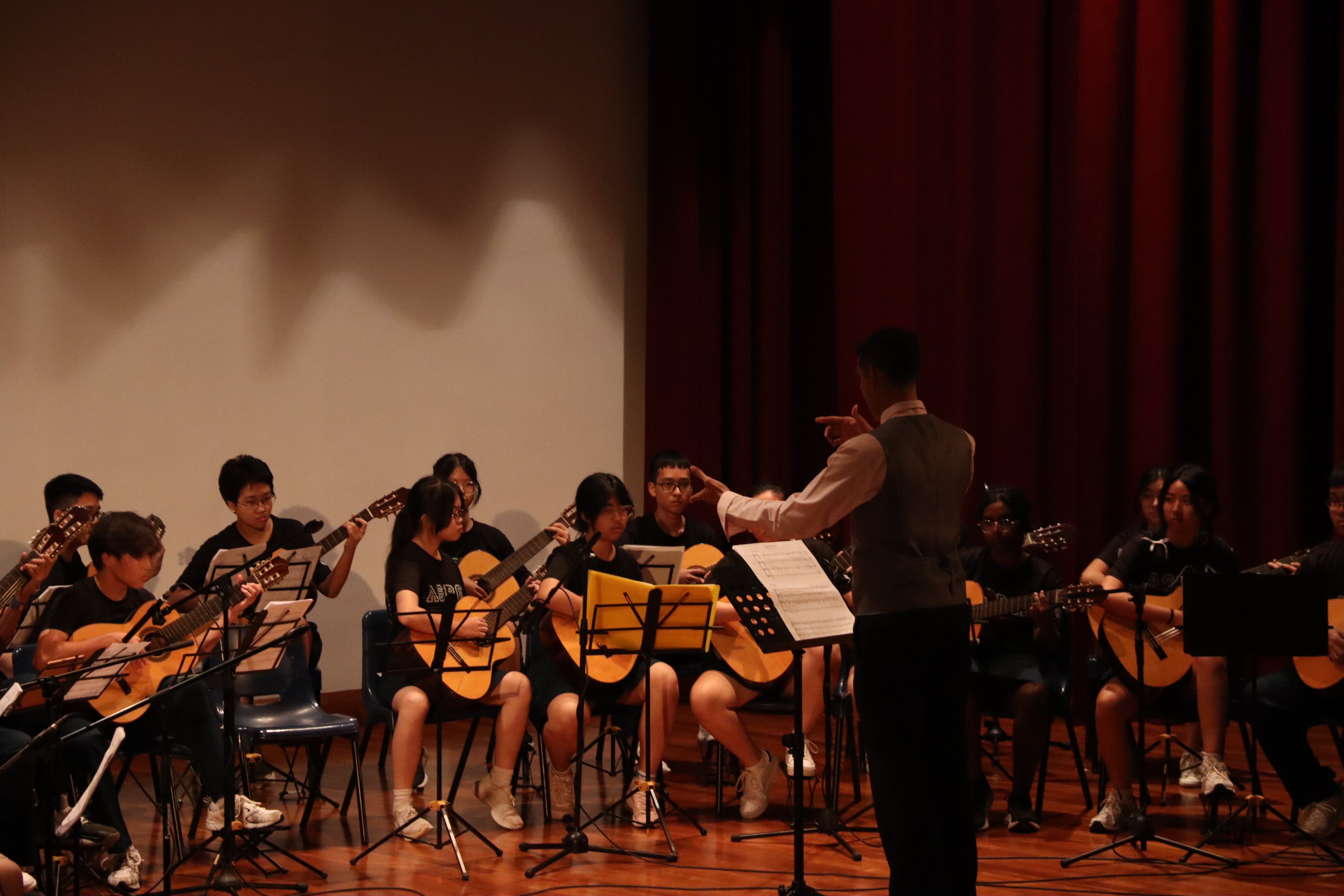 A conductor leads a youth guitar ensemble on stage, with players seated holding acoustic guitars and reading sheet music.