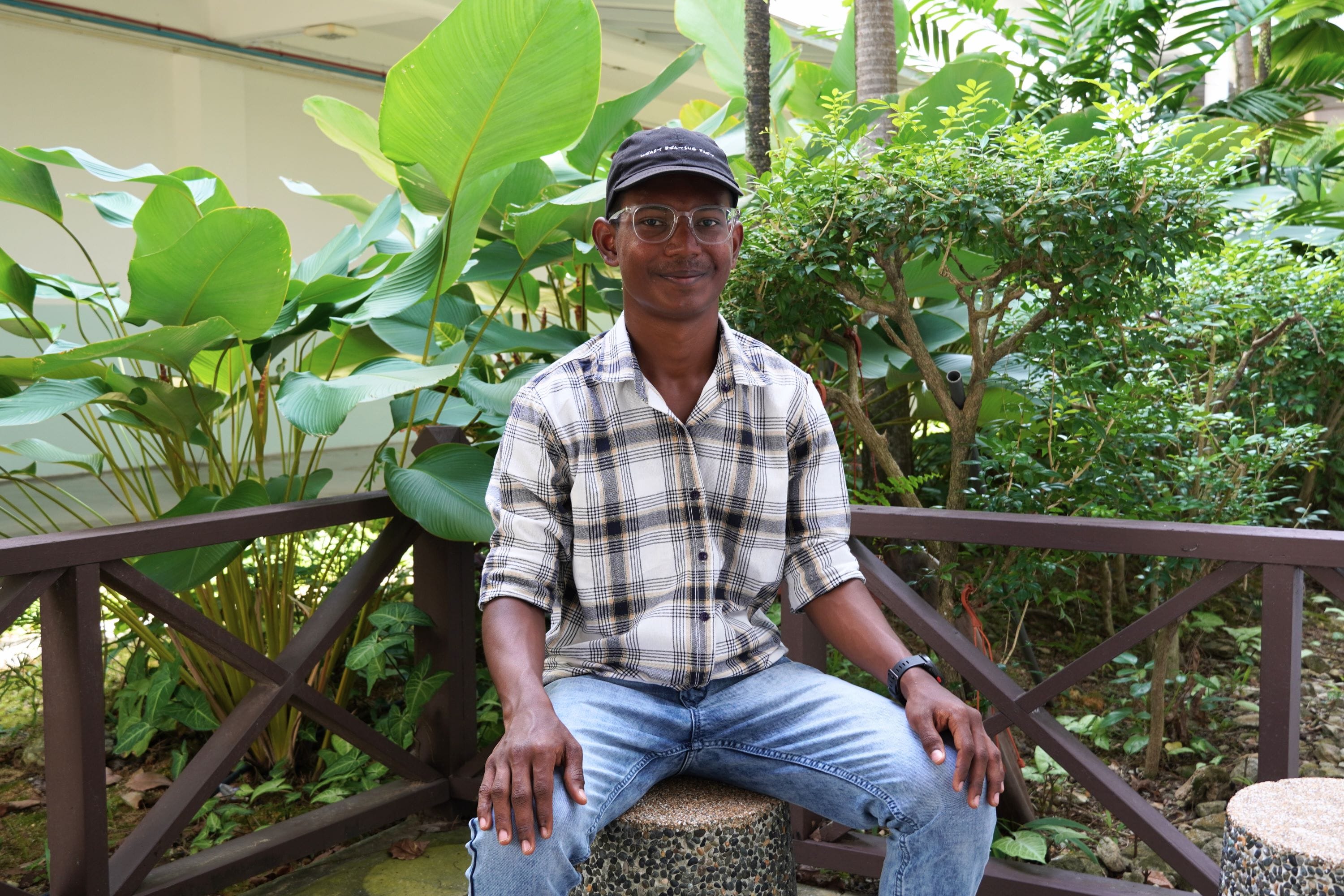 A man wearing glasses, a dark cap, plaid shirt, and jeans sits on a stool in a lush green garden.