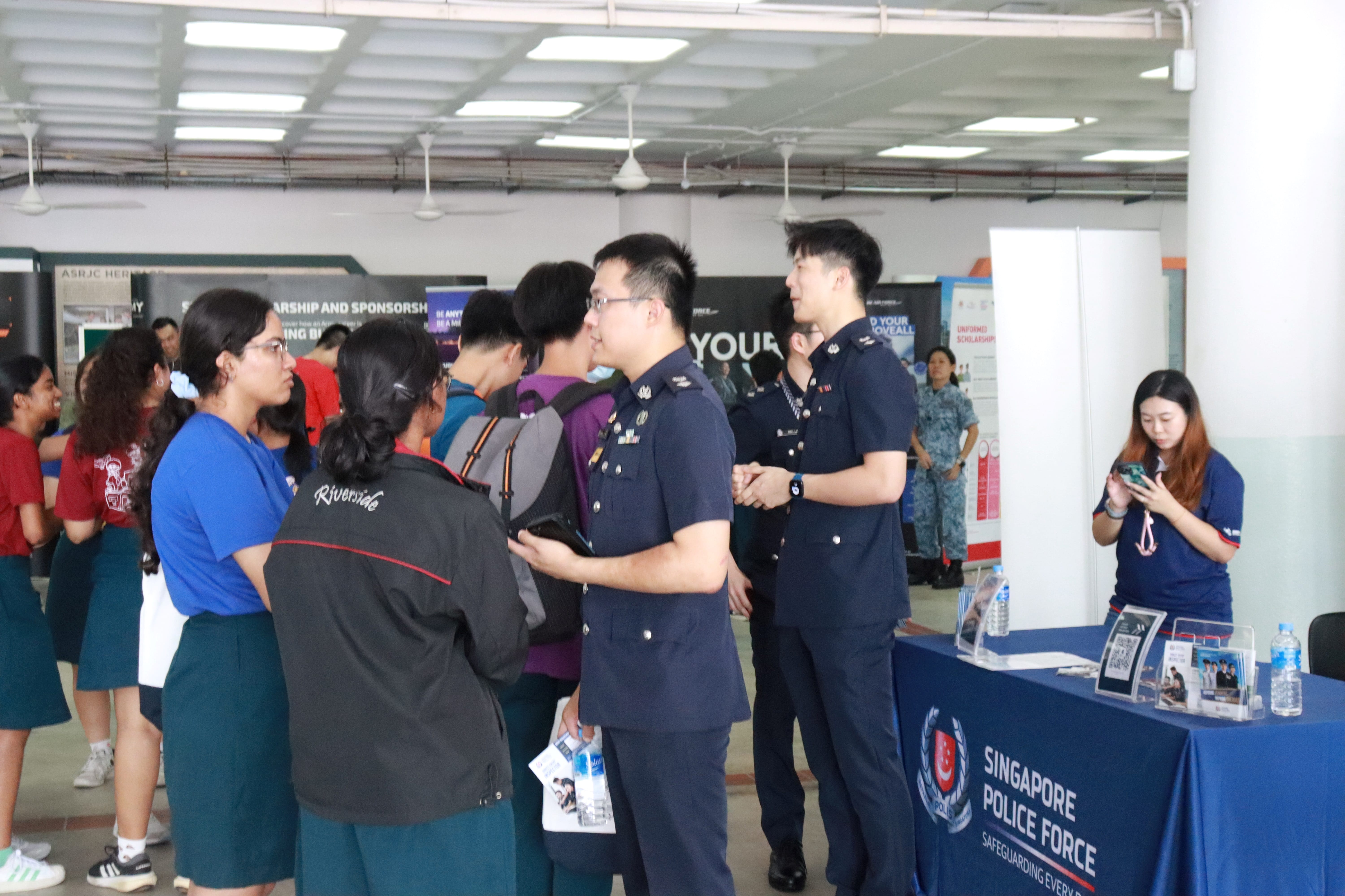 Police officers in uniform talk with students at a booth labeled Singapore Police Force while others mill around.