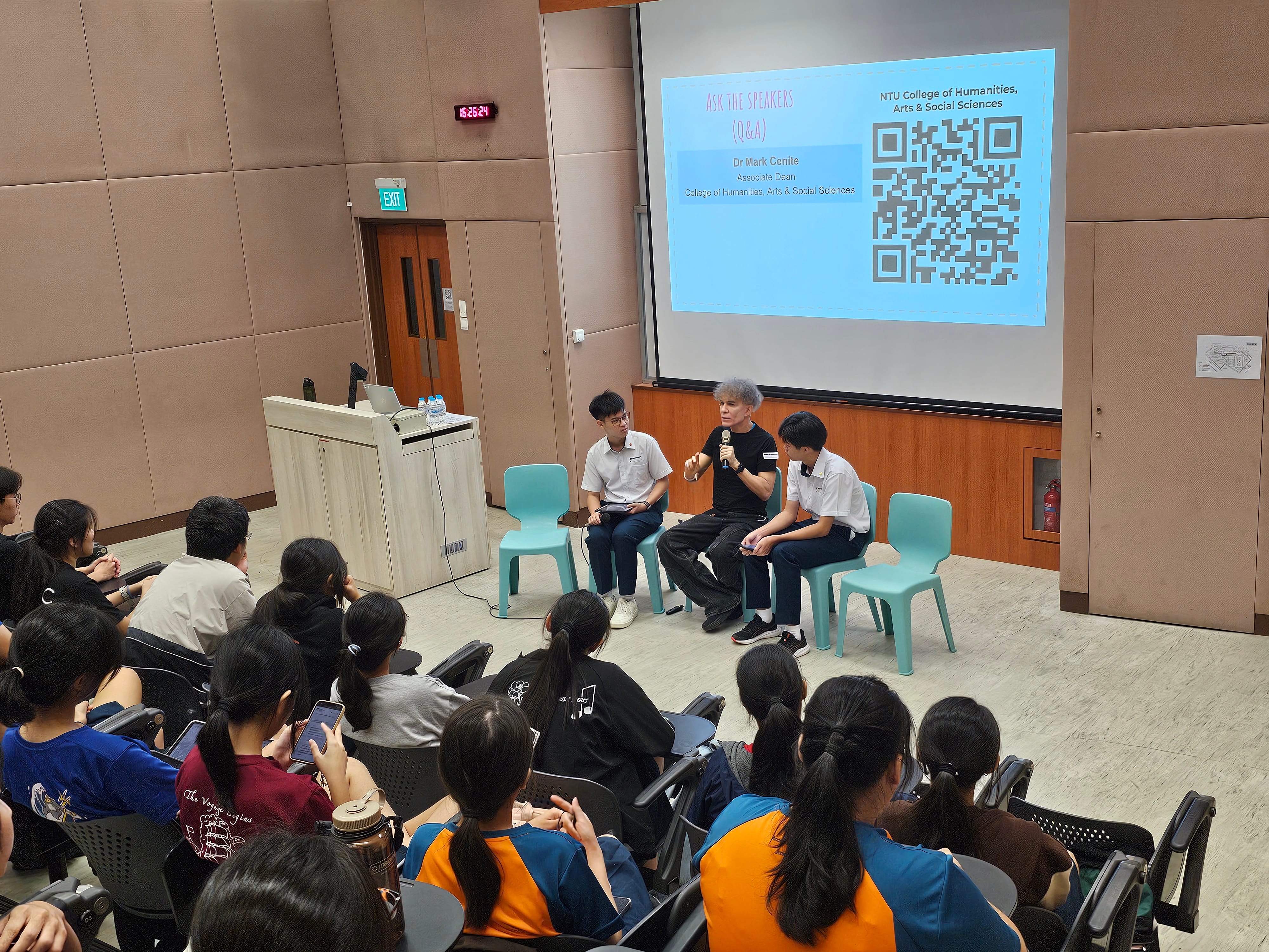Man with microphone talks between two students on chairs as audience watches; slide with QR code projected.