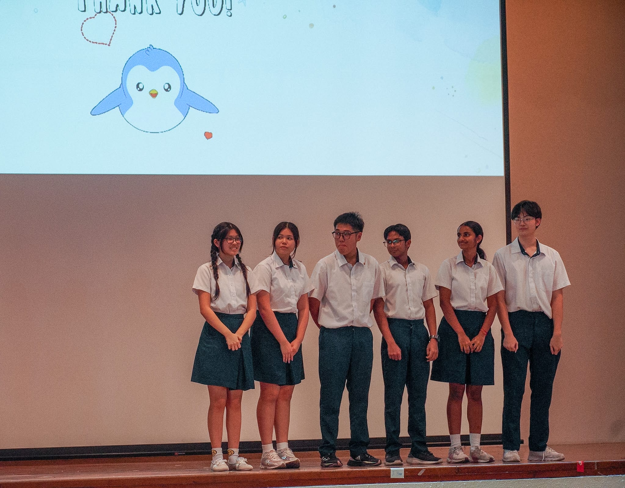 Six students in school uniforms stand on a stage before a projected Thank You slide with a cartoon penguin.