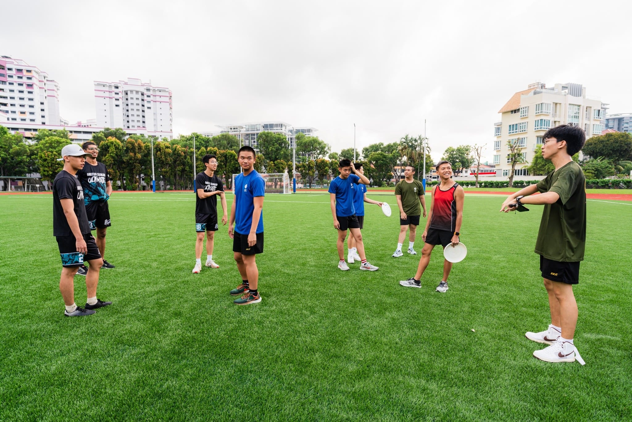 A group of young people holding frisbees stand on a green field listening to a male student giving instructions during an ultimate frisbee session.