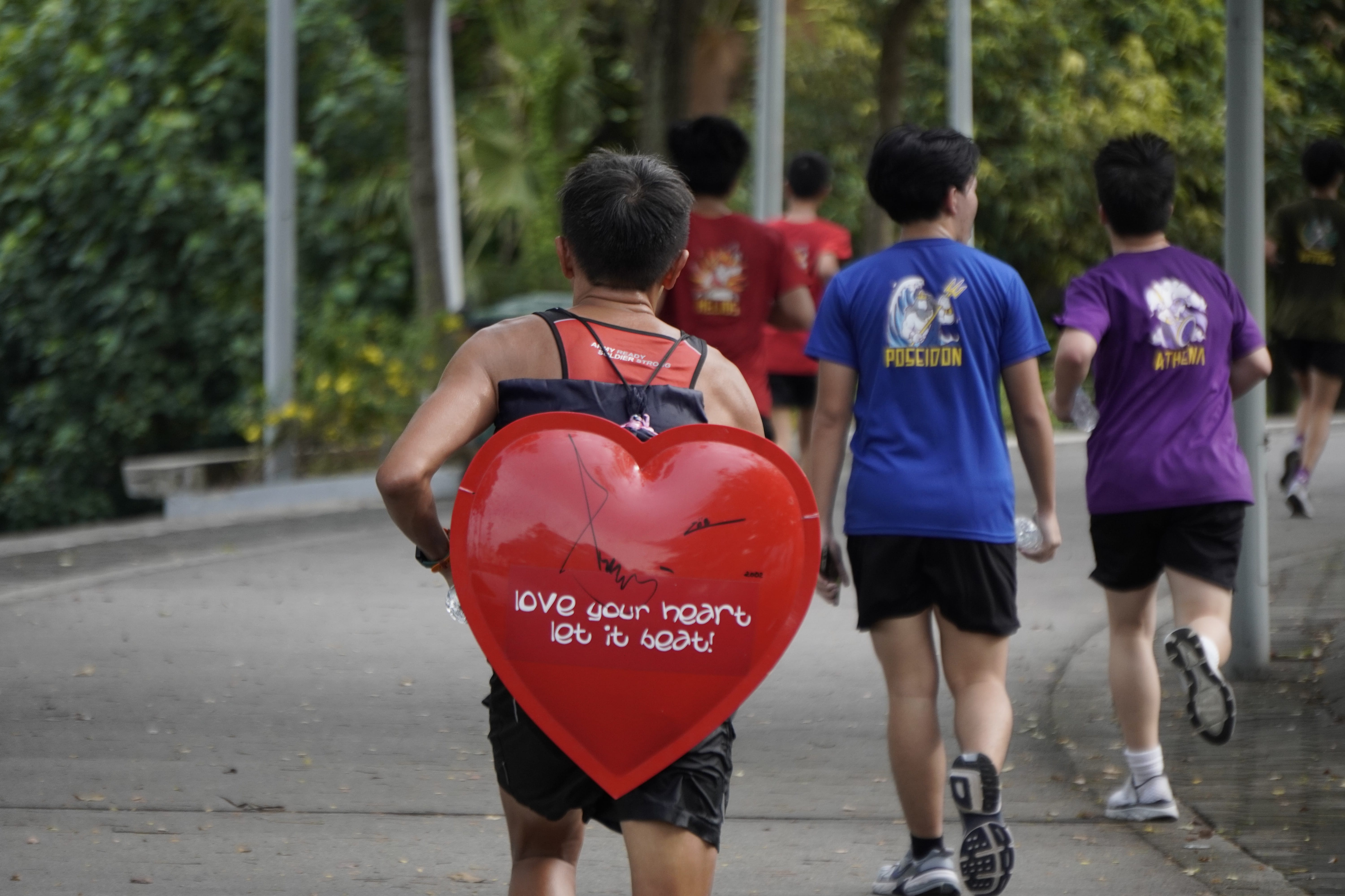 Runner wears a large red heart sign reading love your heart let it beat, with others jogging ahead on a path