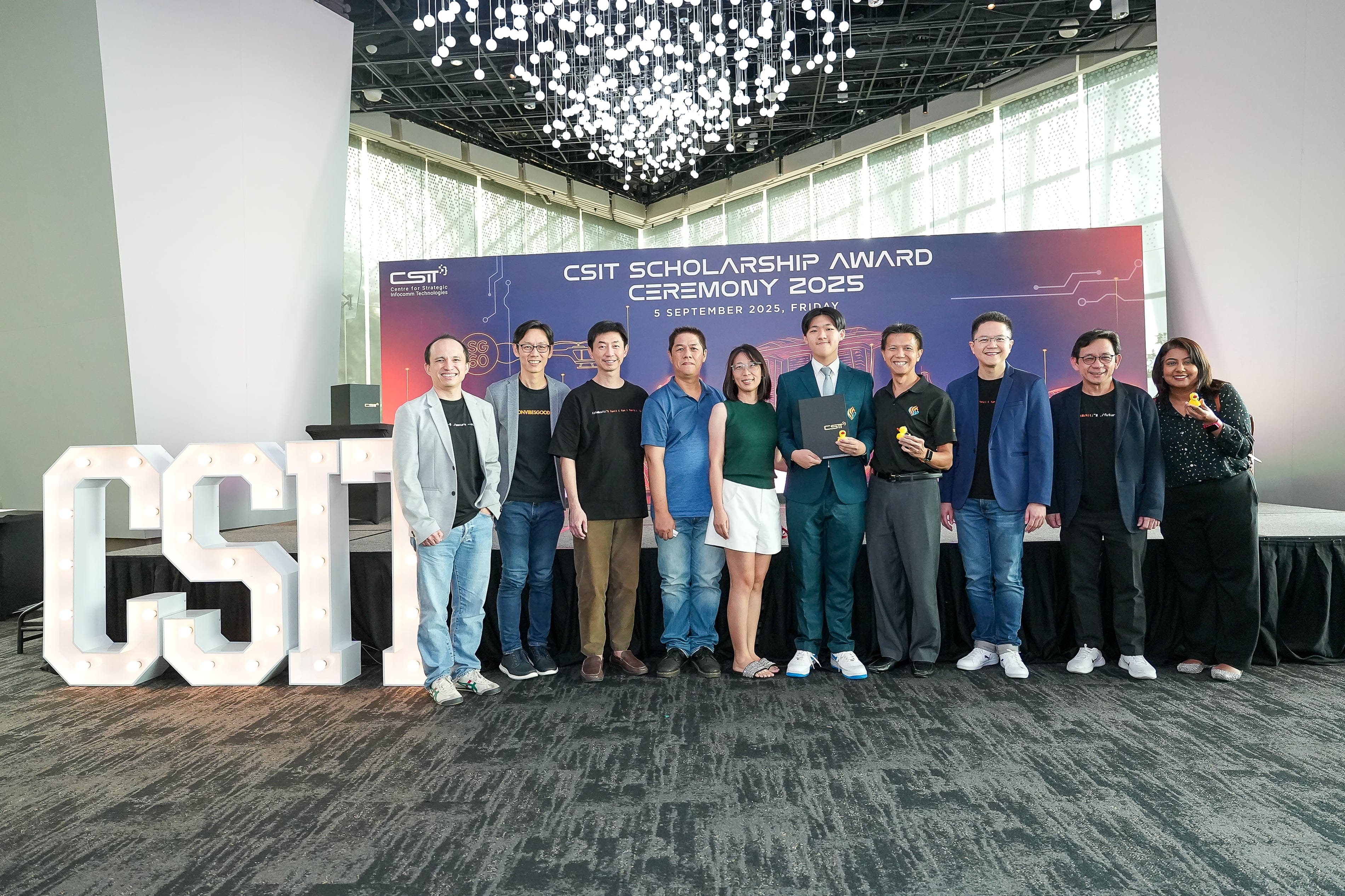 A group of ten people pose at the CSIT Scholarship Award Ceremony 2025 banner, with lit CSIT letters nearby.