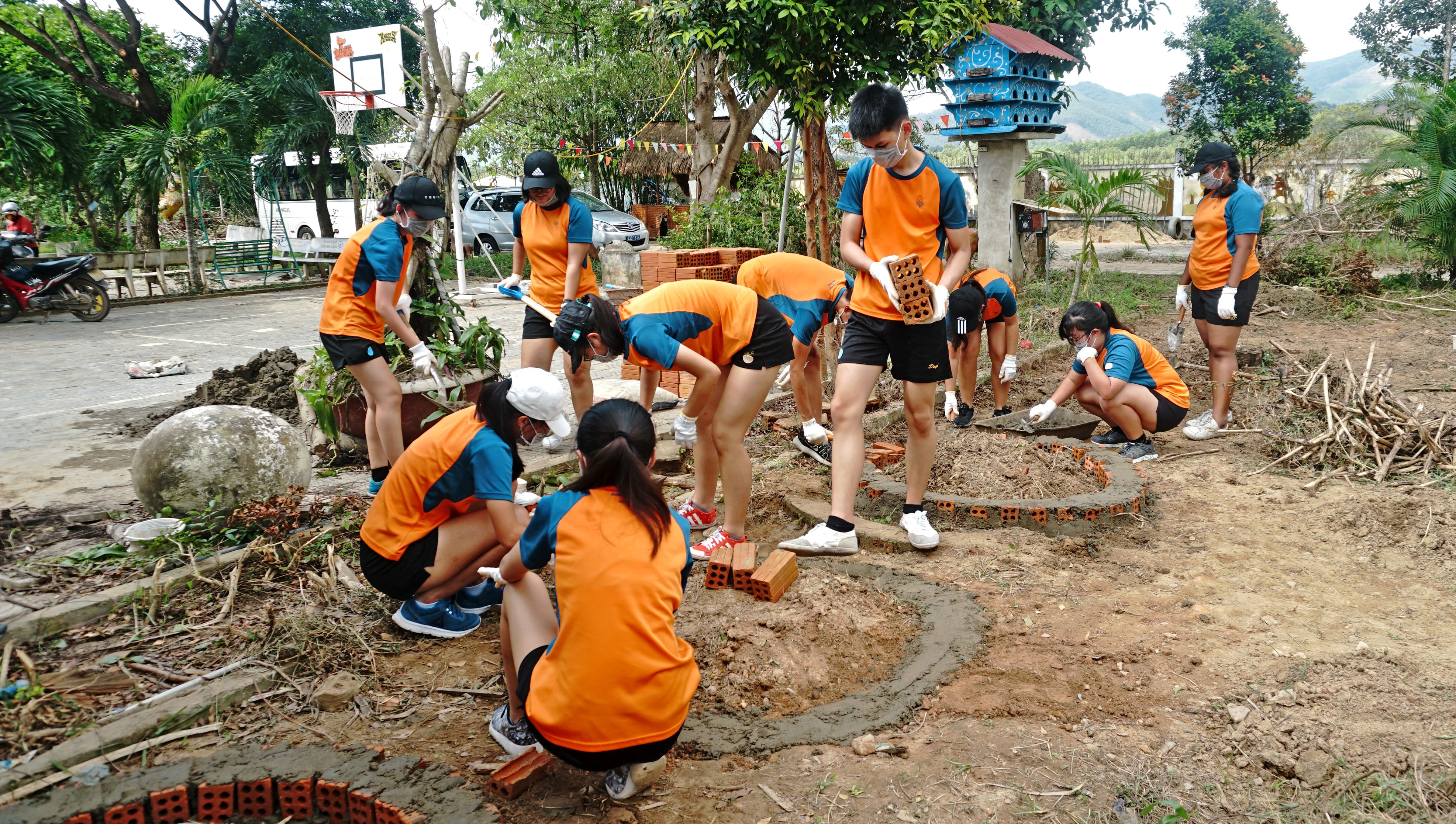 Group in orange shirts and masks lay bricks and spread cement outdoors near trees and a basketball hoop.