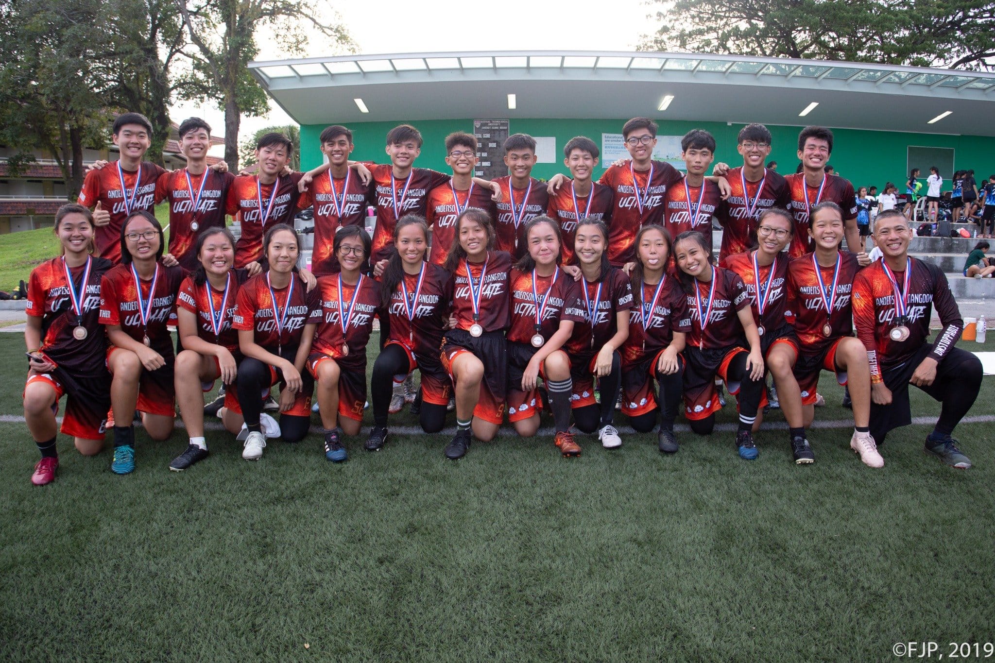 A large youth sports team in red uniforms with medals poses together on a grass field outdoors.