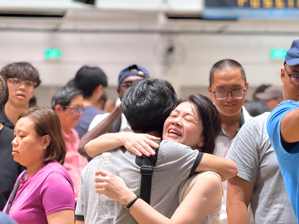 A smiling woman hugs a person tightly in a crowd of people inside a building.