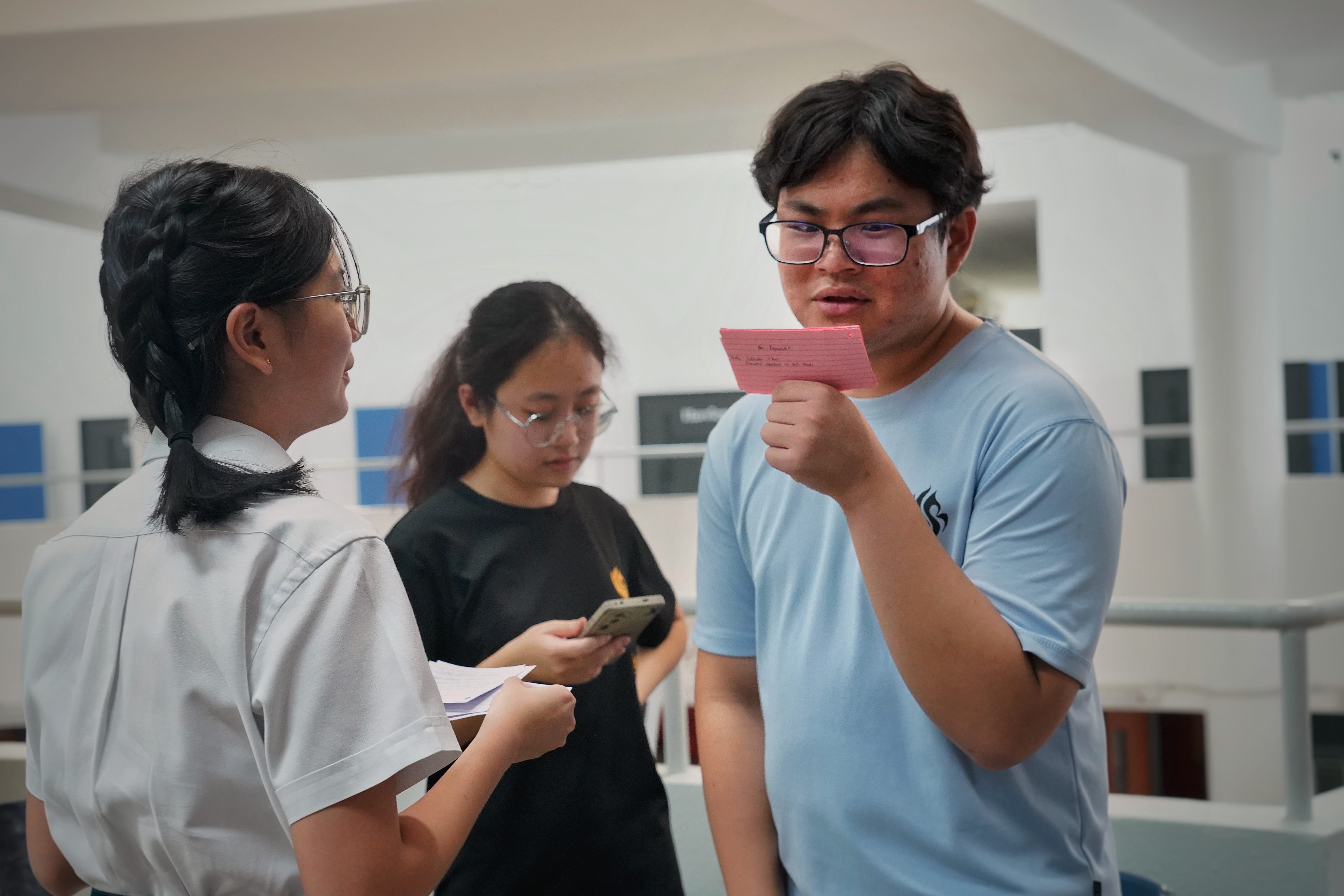 Young man in glasses reads a pink card while two young women nearby hold papers and a phone indoors.