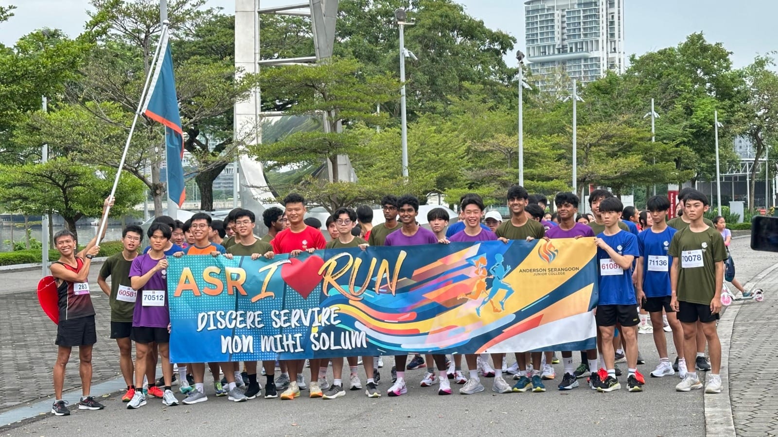 A group of students with race bibs hold an Anderson Serangoon Junior College "ASR I Run" banner outdoors.