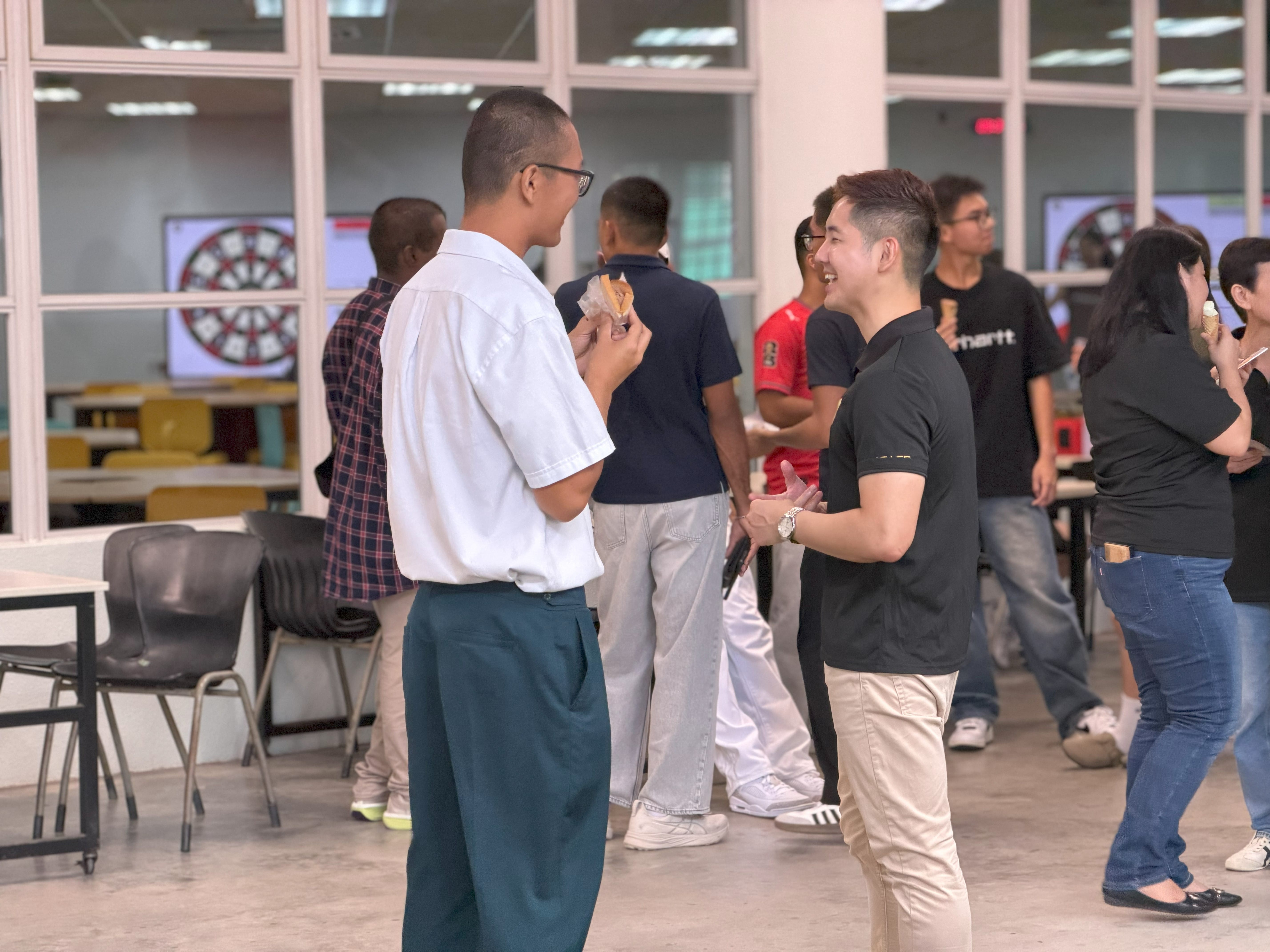 Male student in white shirt, eating a sandwich, talks to a man in black shirt indoors