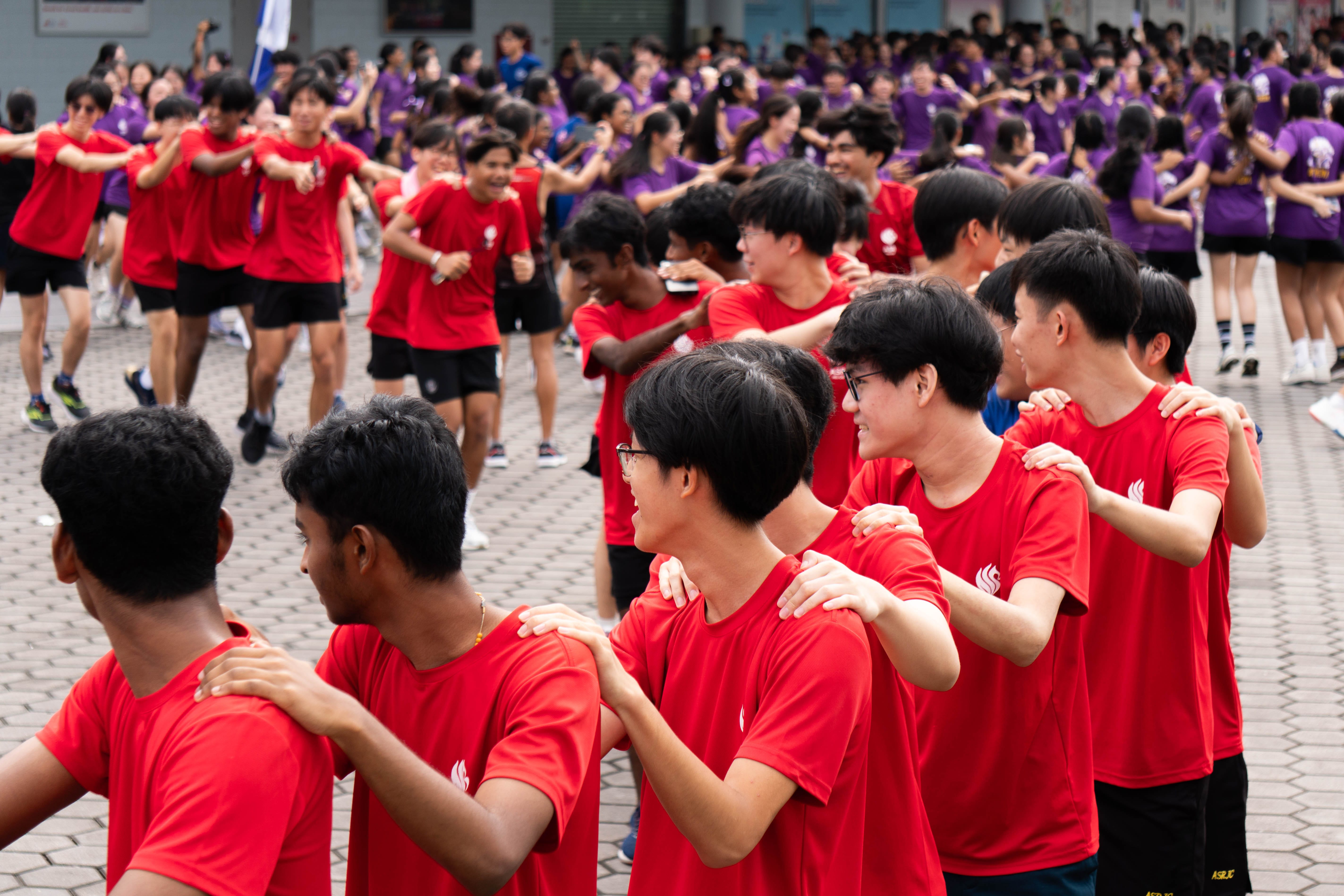 Large group of students in red and purple shirts dancing together outdoors on a paved courtyard.