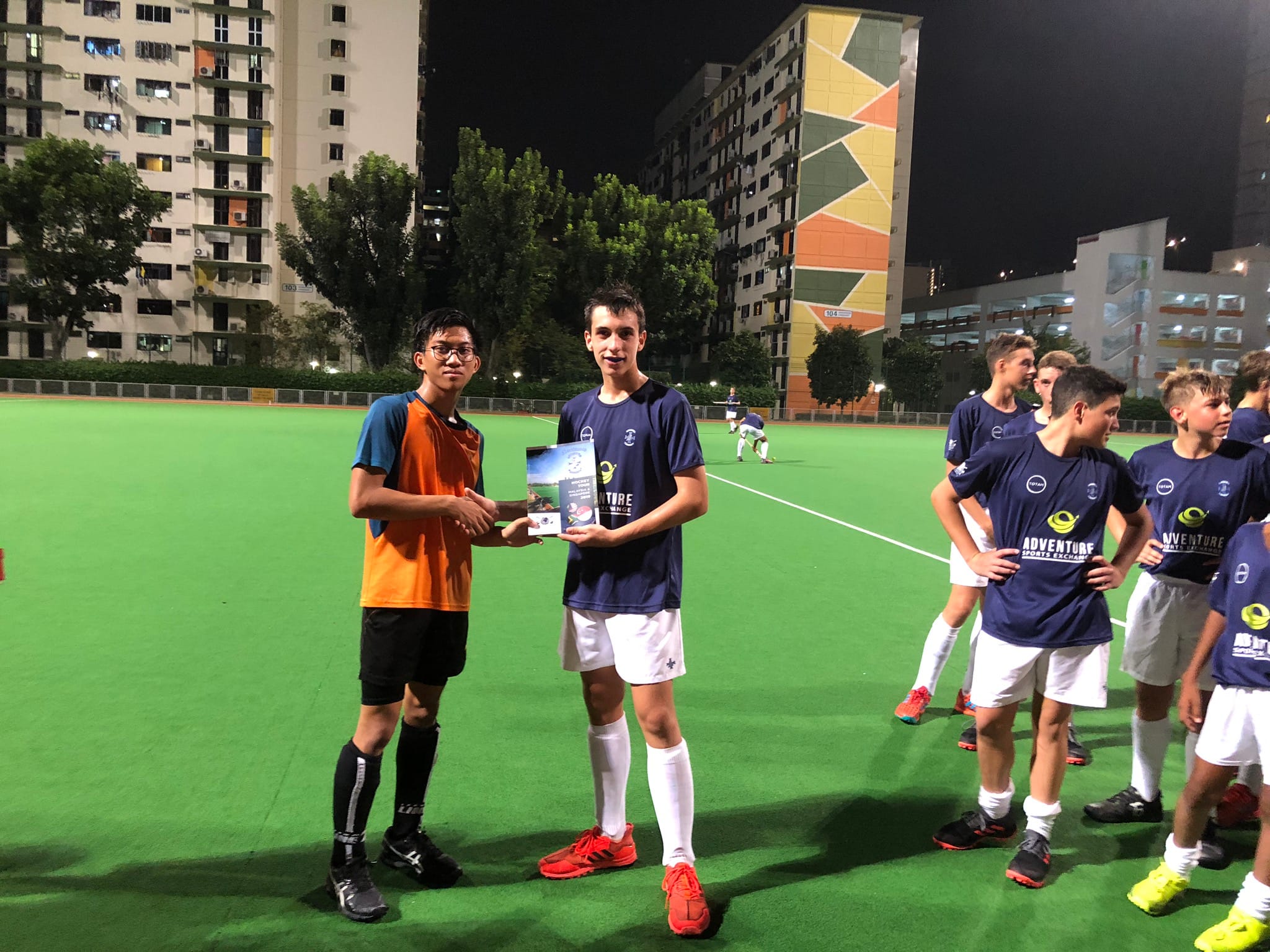 Two boys shake hands on a lit green sports field at night, holding a book, with teammates and buildings behind them.