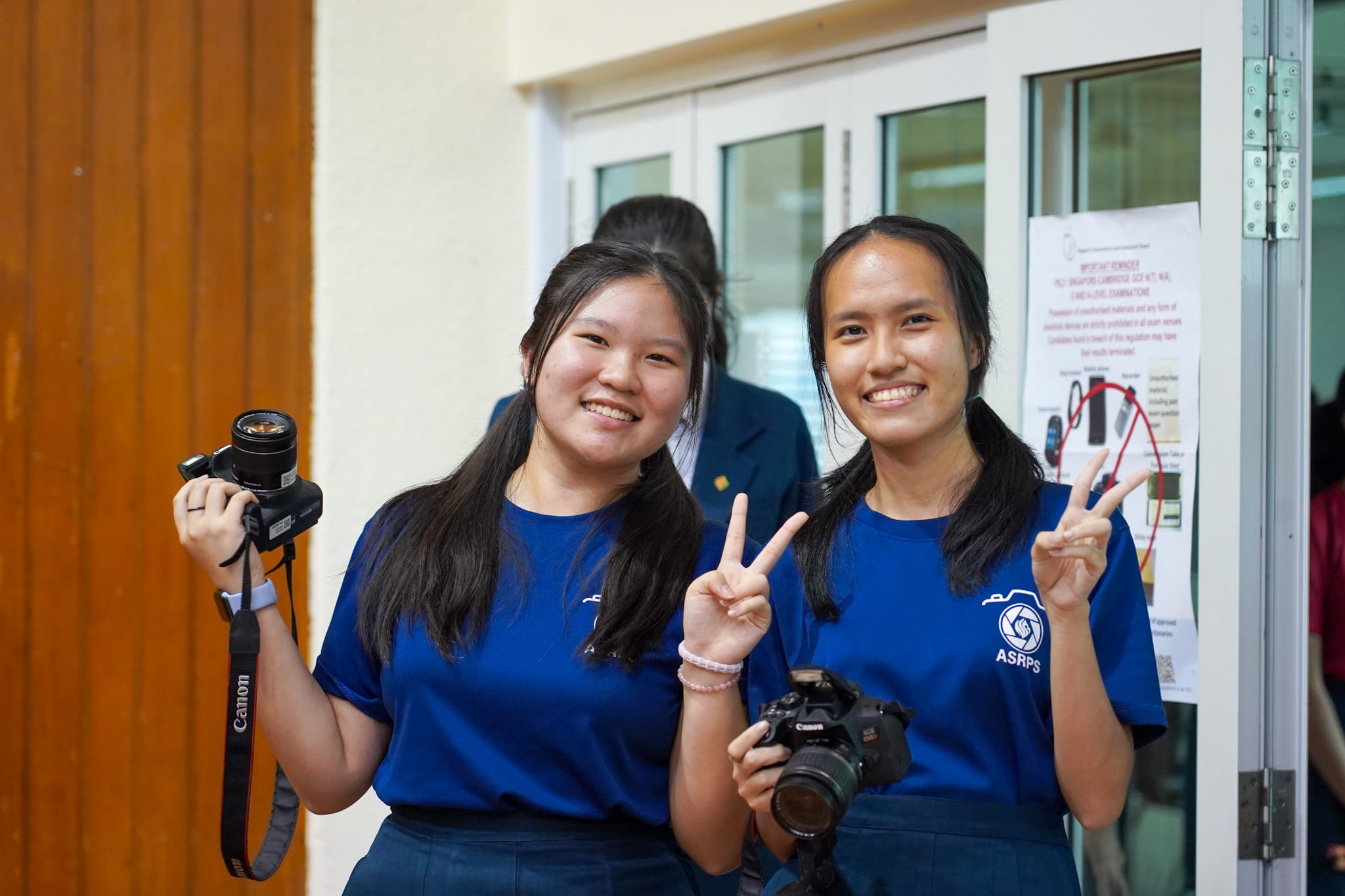 Two smiling girls in blue ASRPS shirts hold Canon cameras and flash peace signs near a glass door.