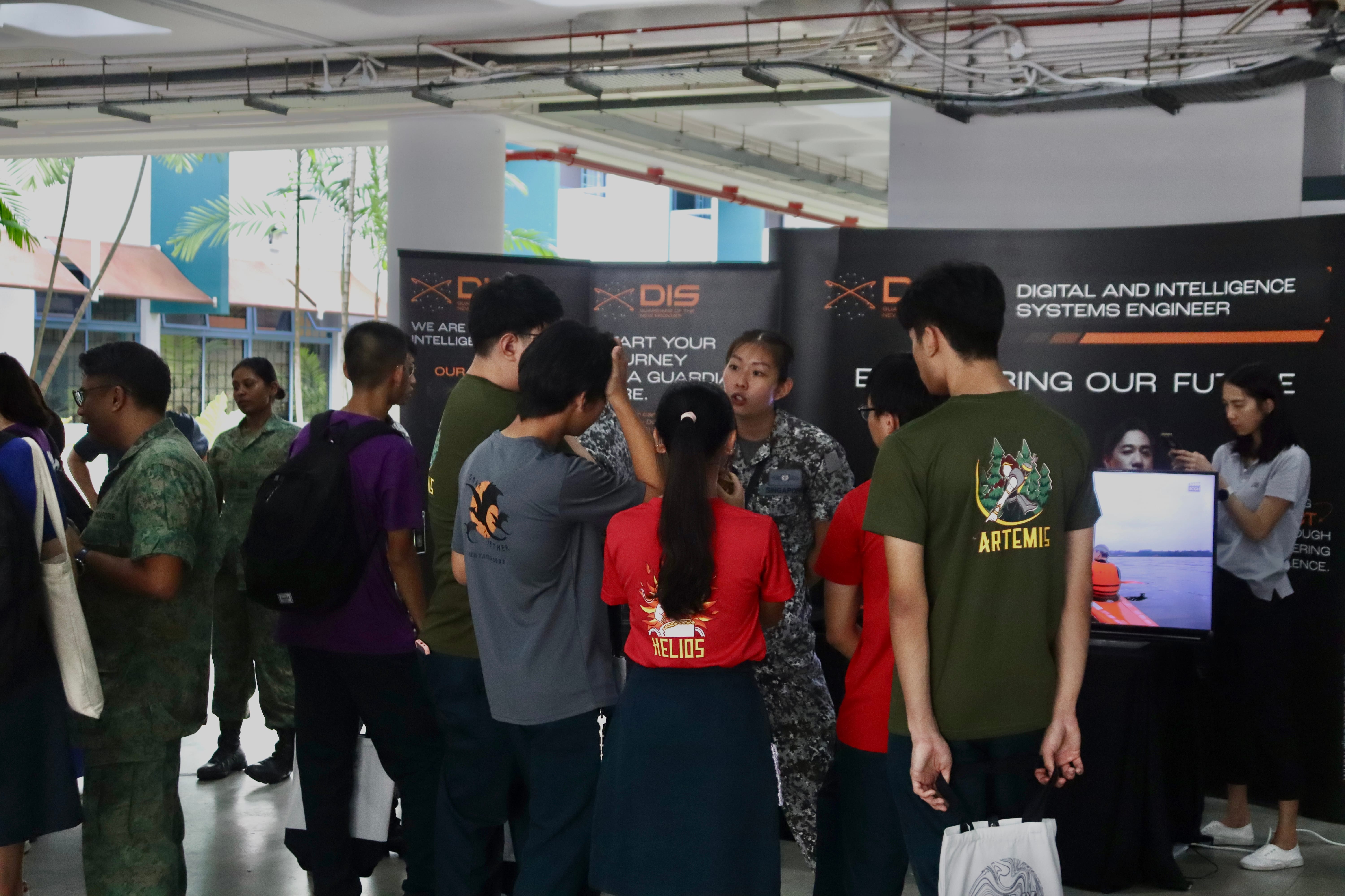 Crowd gathered at an indoor booth with DIS banners, speaking to a person in camouflage uniform near a monitor.