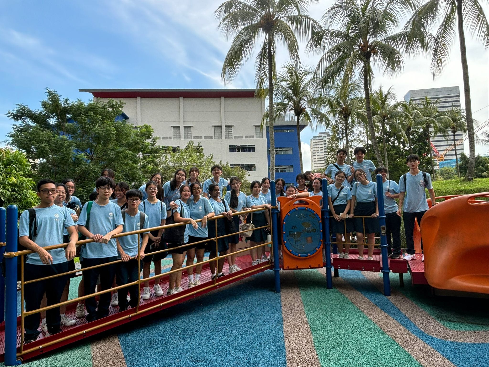 A large group of teenagers in matching light blue shirts pose together on a colorful outdoor playground structure.