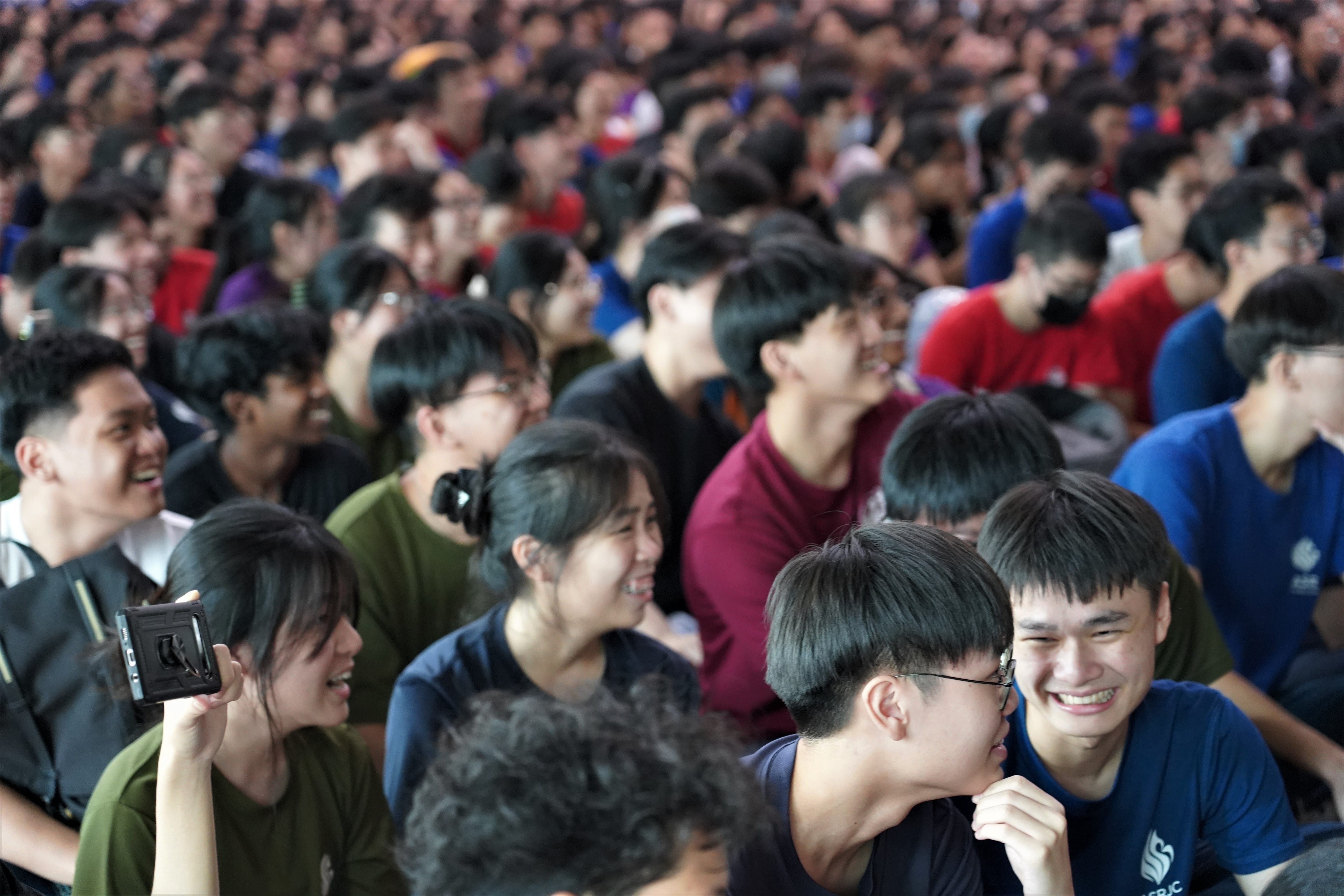 Large crowd seated closely together, many smiling and laughing; one person at left holds up a phone.