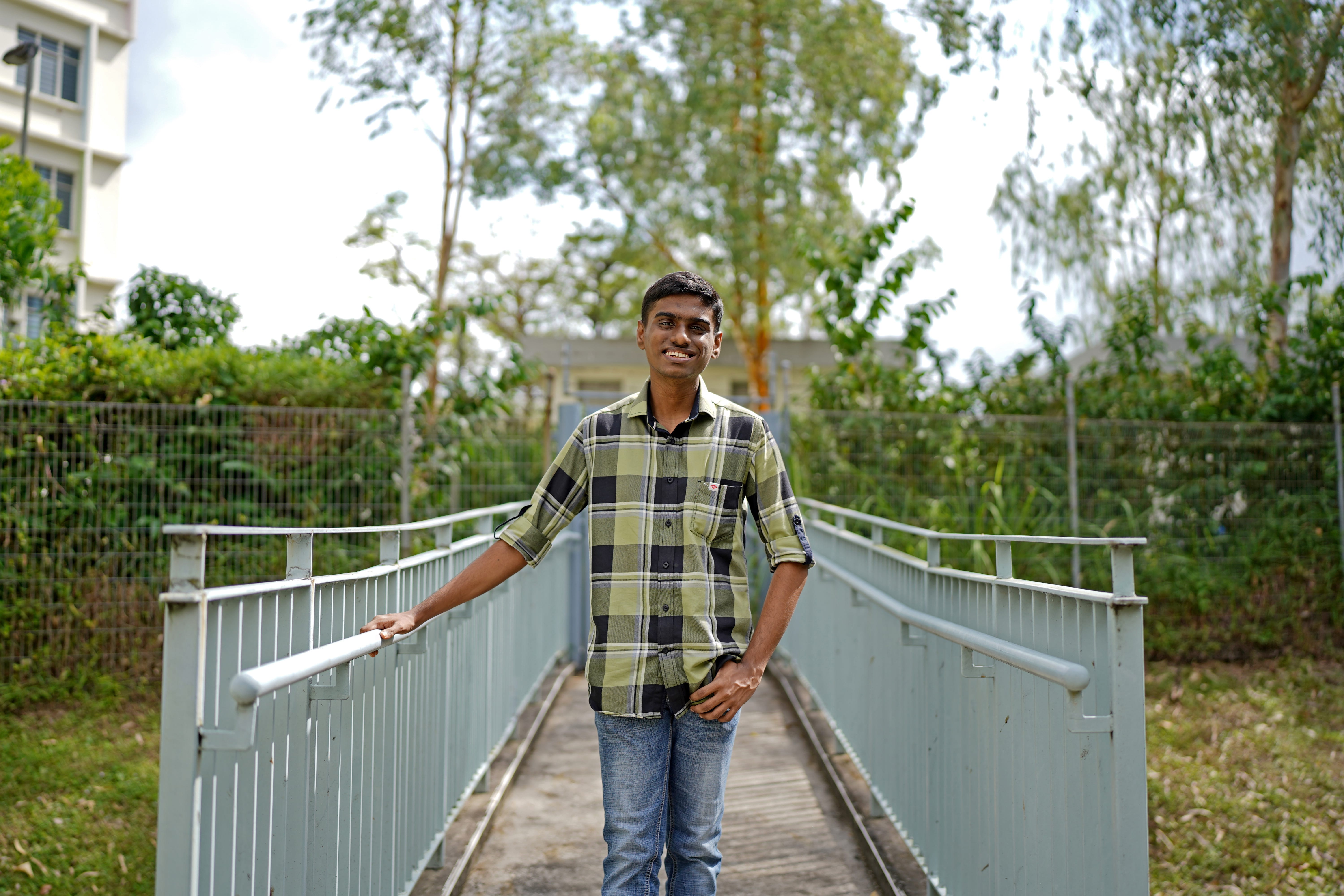 Smiling man in a plaid shirt stands on a narrow metal footbridge, holding the rail, with trees behind.
