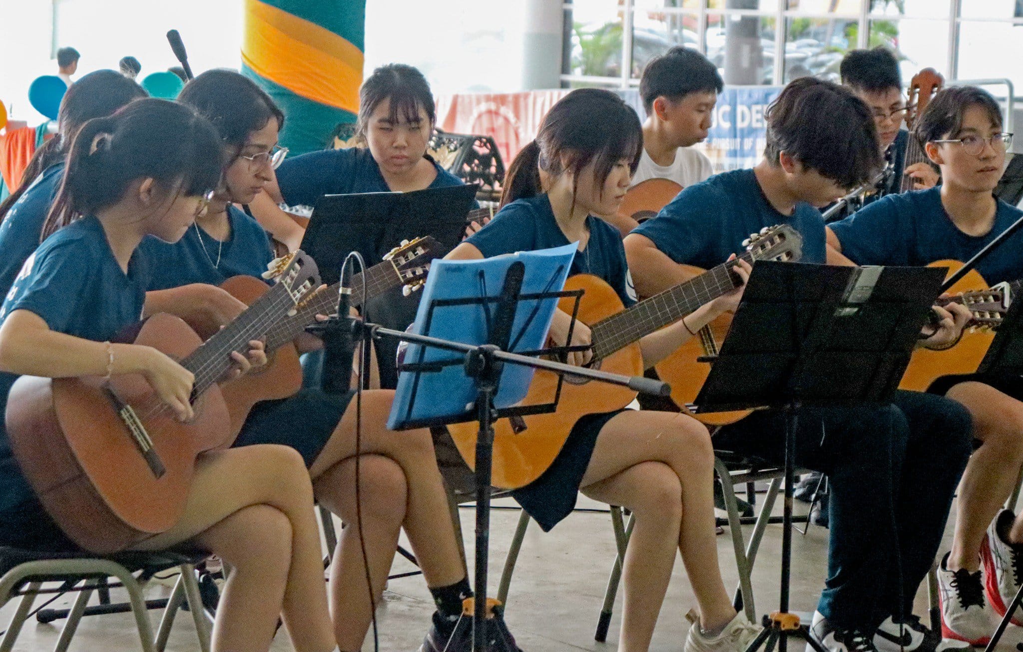A group of young people in blue shirts sit playing acoustic guitars with music stands in front of them.