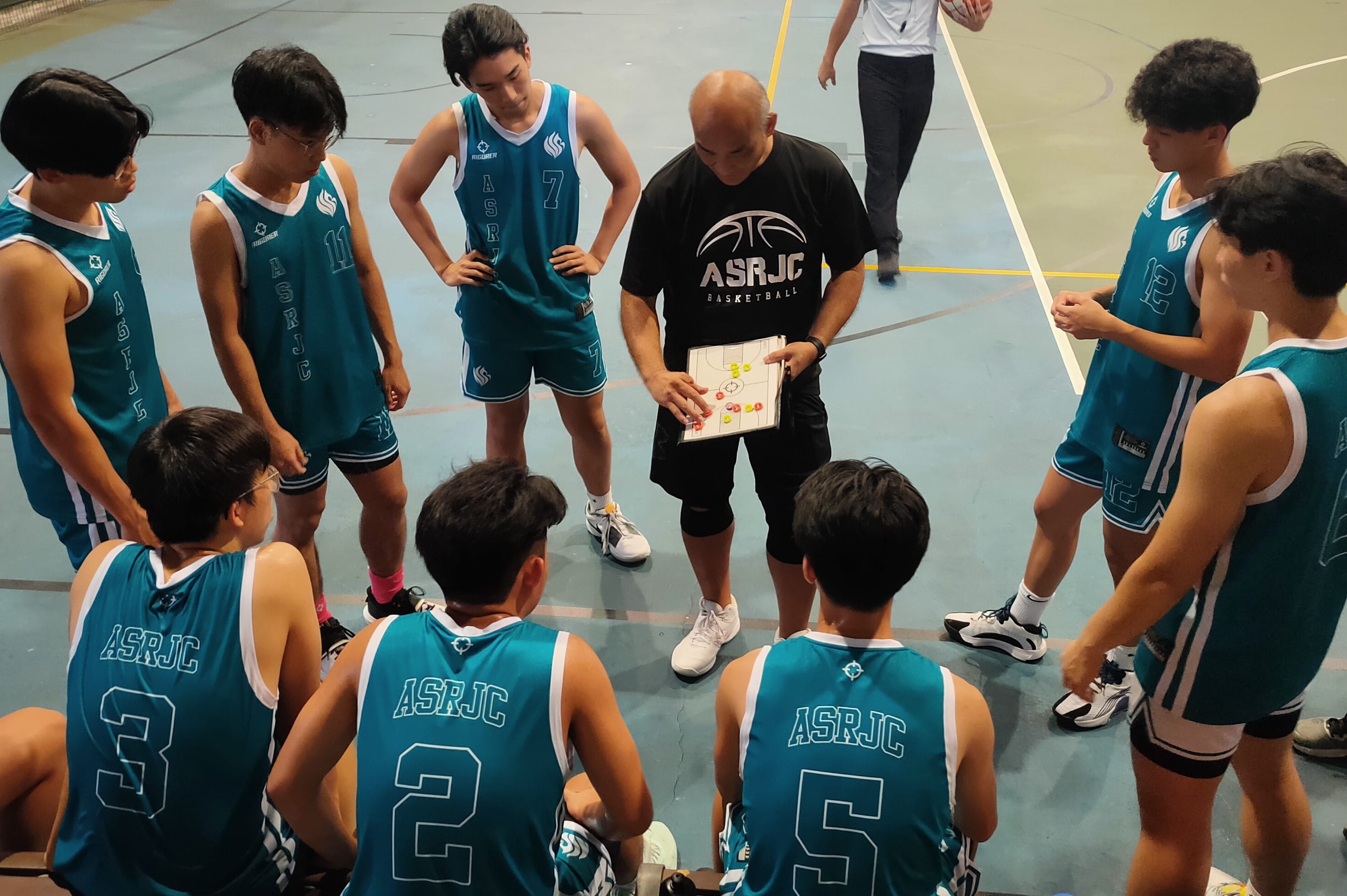 A coach in a black ASRJC shirt shows a play diagram to young basketball players in teal ASRJC jerseys.