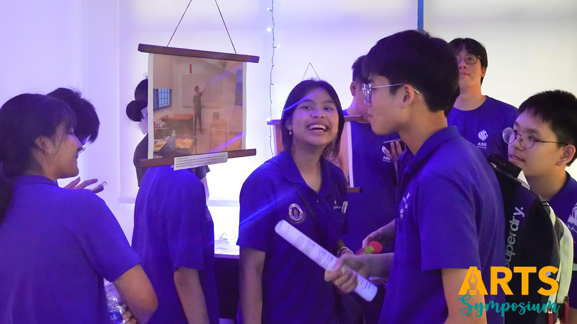 Students in purple uniforms laugh and chat at an Arts Symposium event near a hanging artwork display.