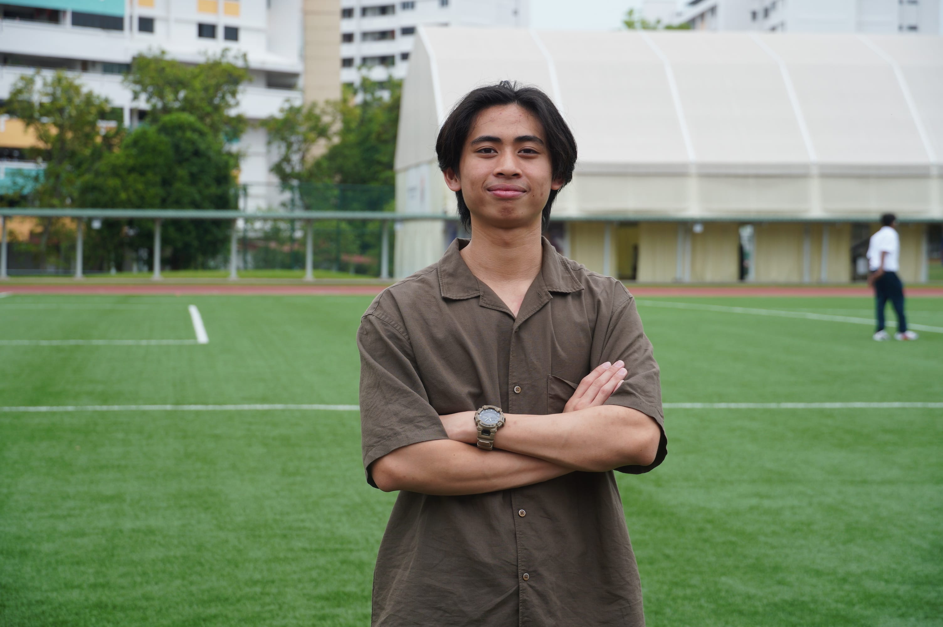 Young man with crossed arms and a watch, smiling on a green sports field with buildings in the background.