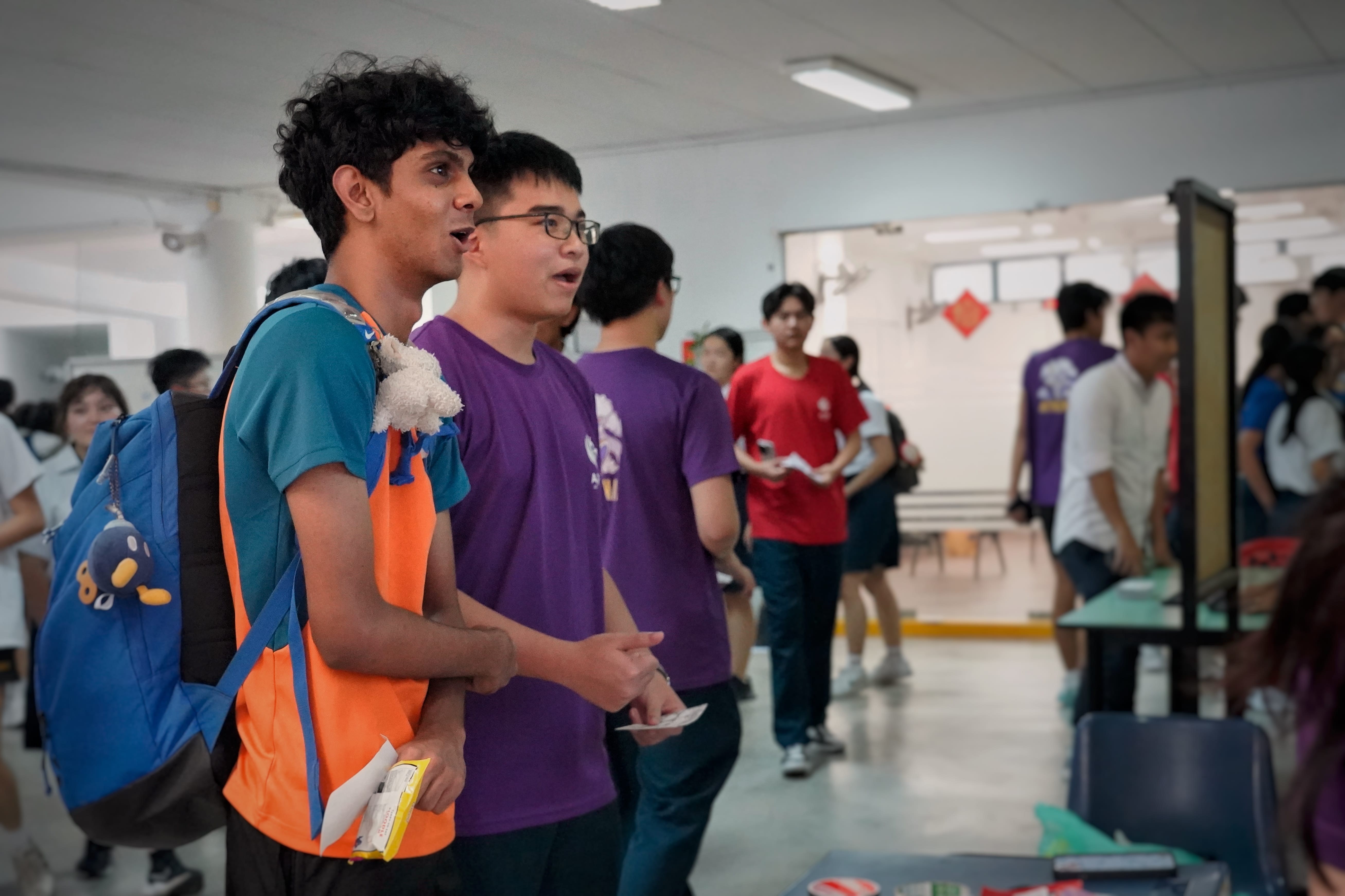 Two young men stand in a busy indoor hall, one with a blue backpack and orange vest, the other in a purple shirt.