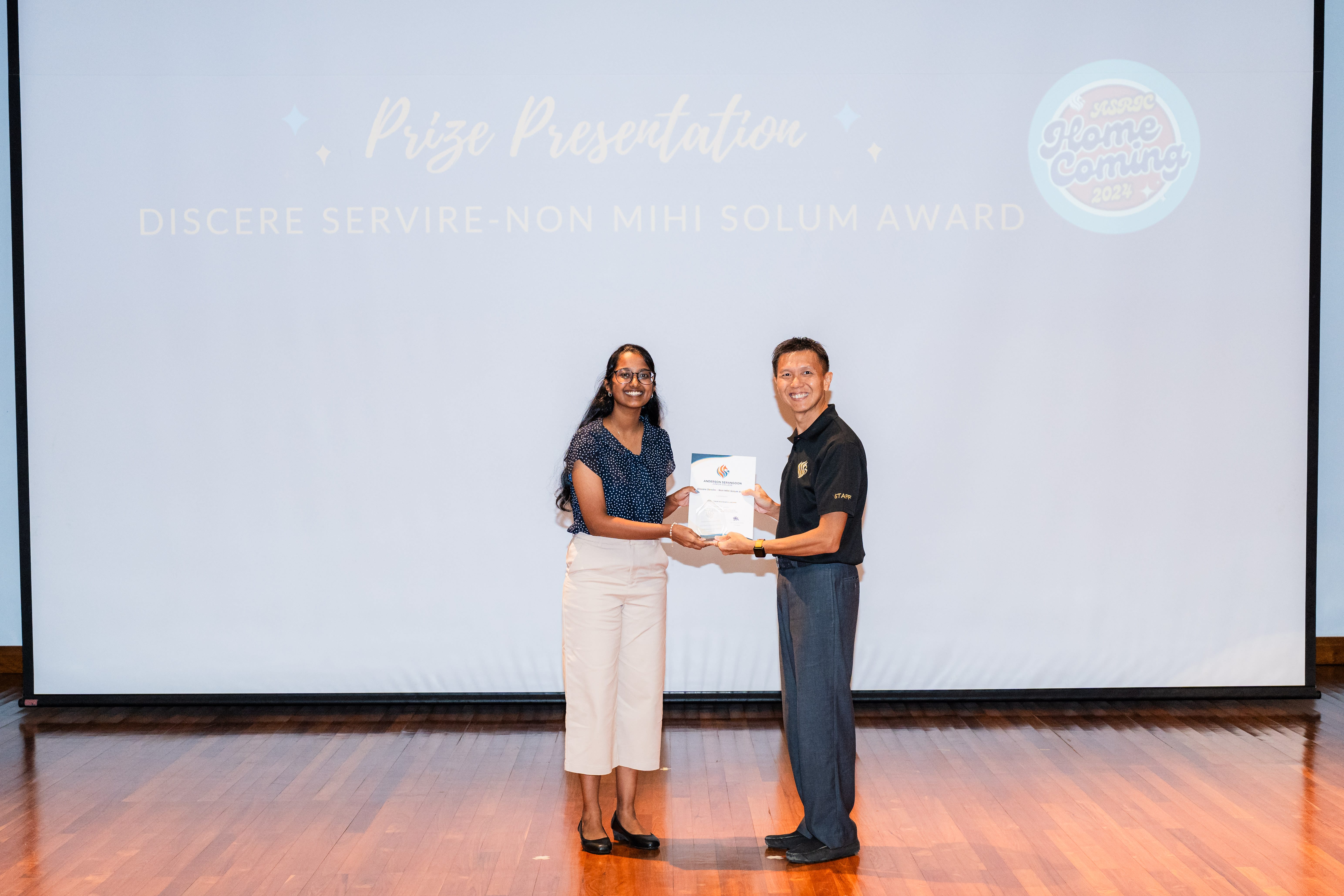 Two people stand on a stage holding a certificate in front of a Prize Presentation screen for the Discere Servire Non Mihi Solum Award.
