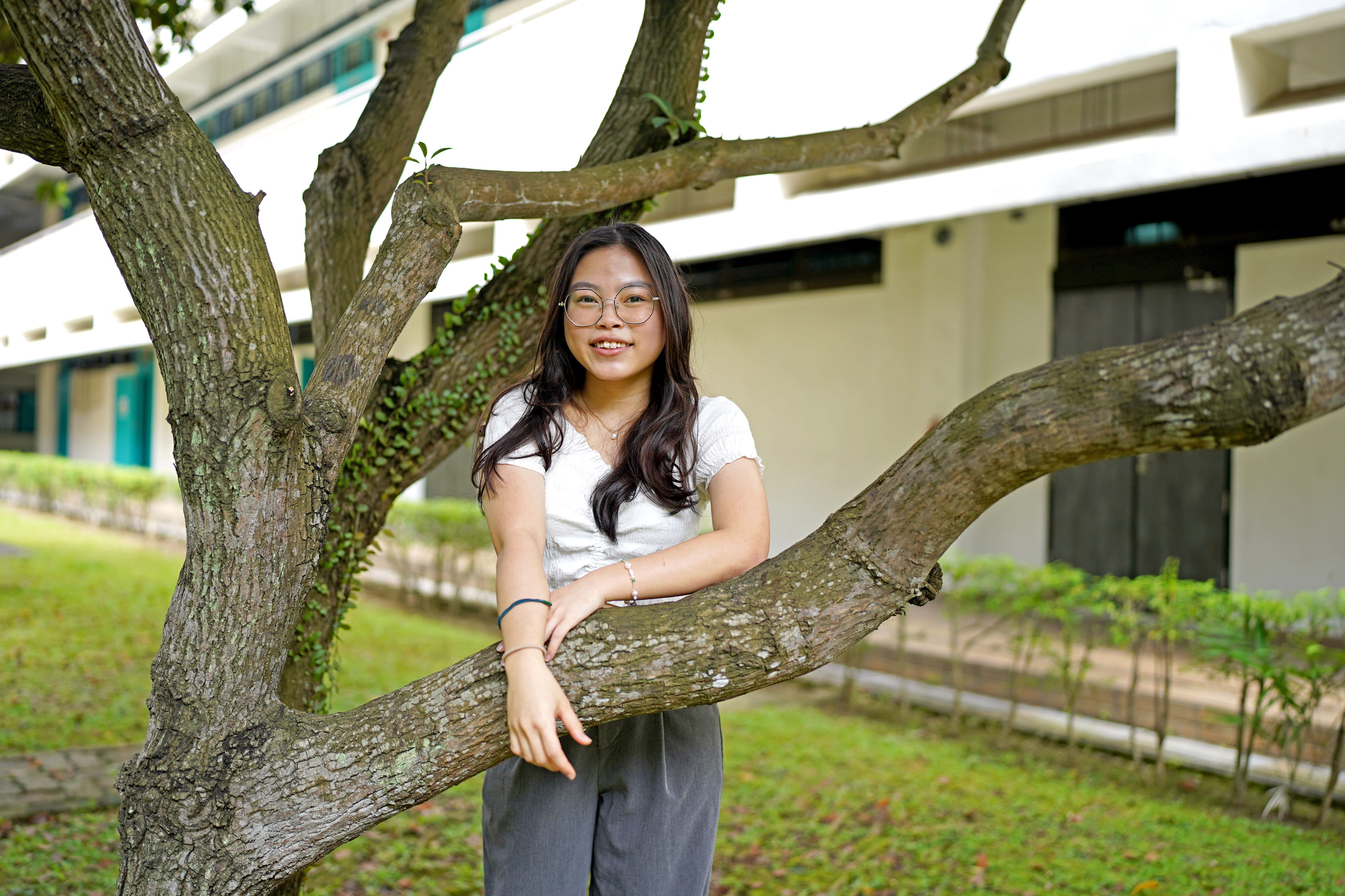 Young woman with glasses leans on a large tree branch, smiling, with a building and grass behind.