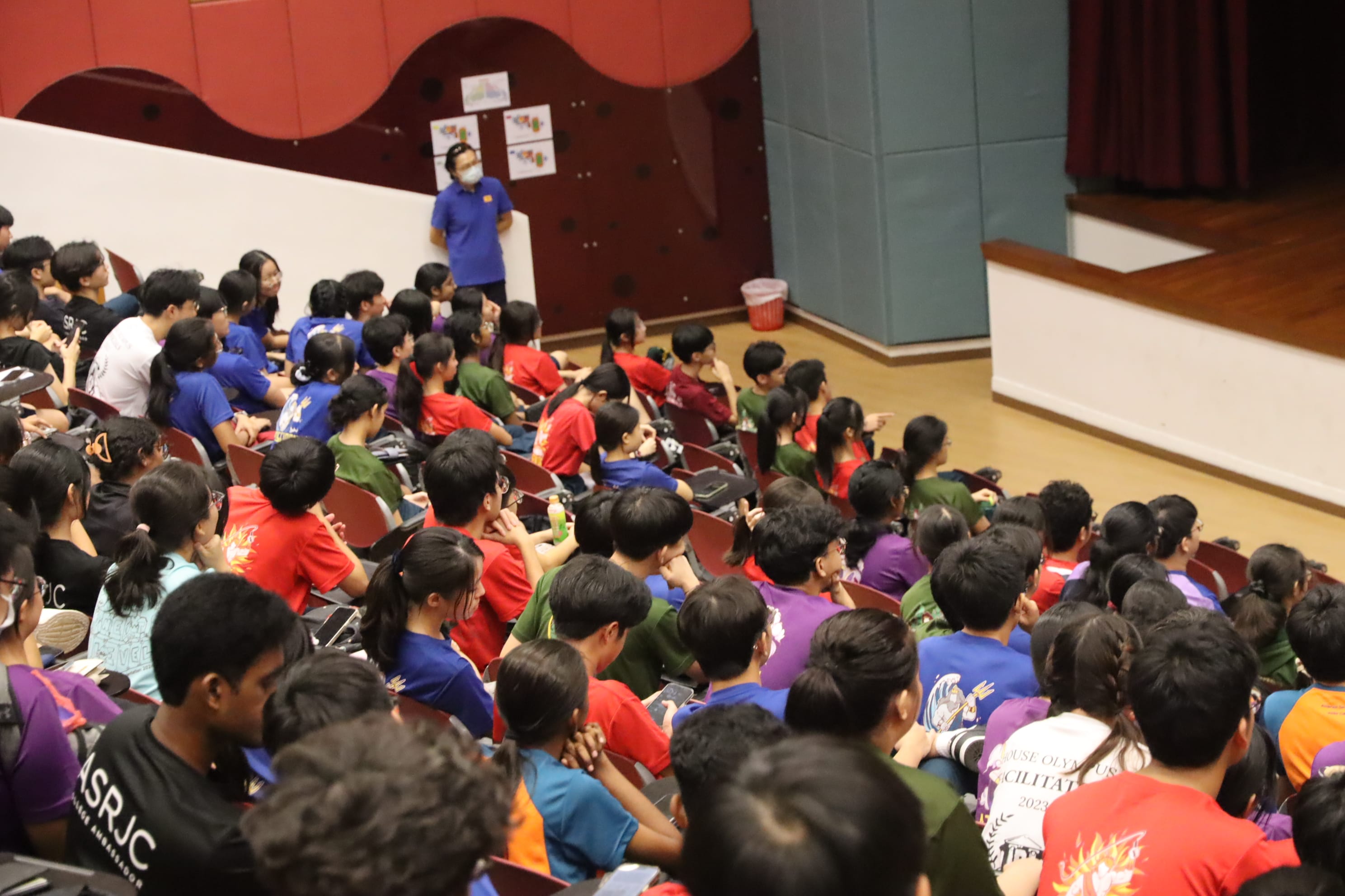 A large group of students in colorful shirts sit in an auditorium facing a masked speaker at a podium.