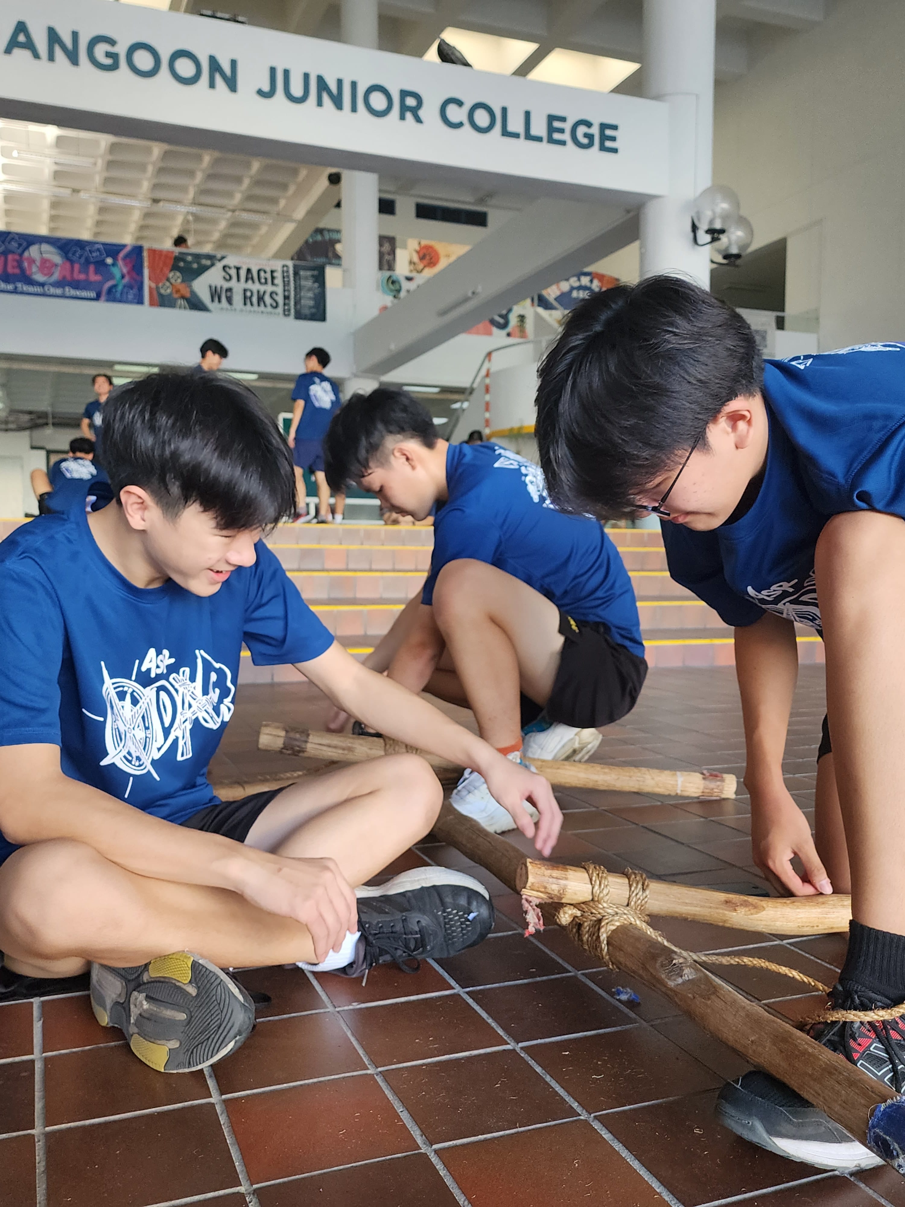 Students in blue shirts assemble wooden poles tied with rope on a tiled floor at Angoon Junior College.