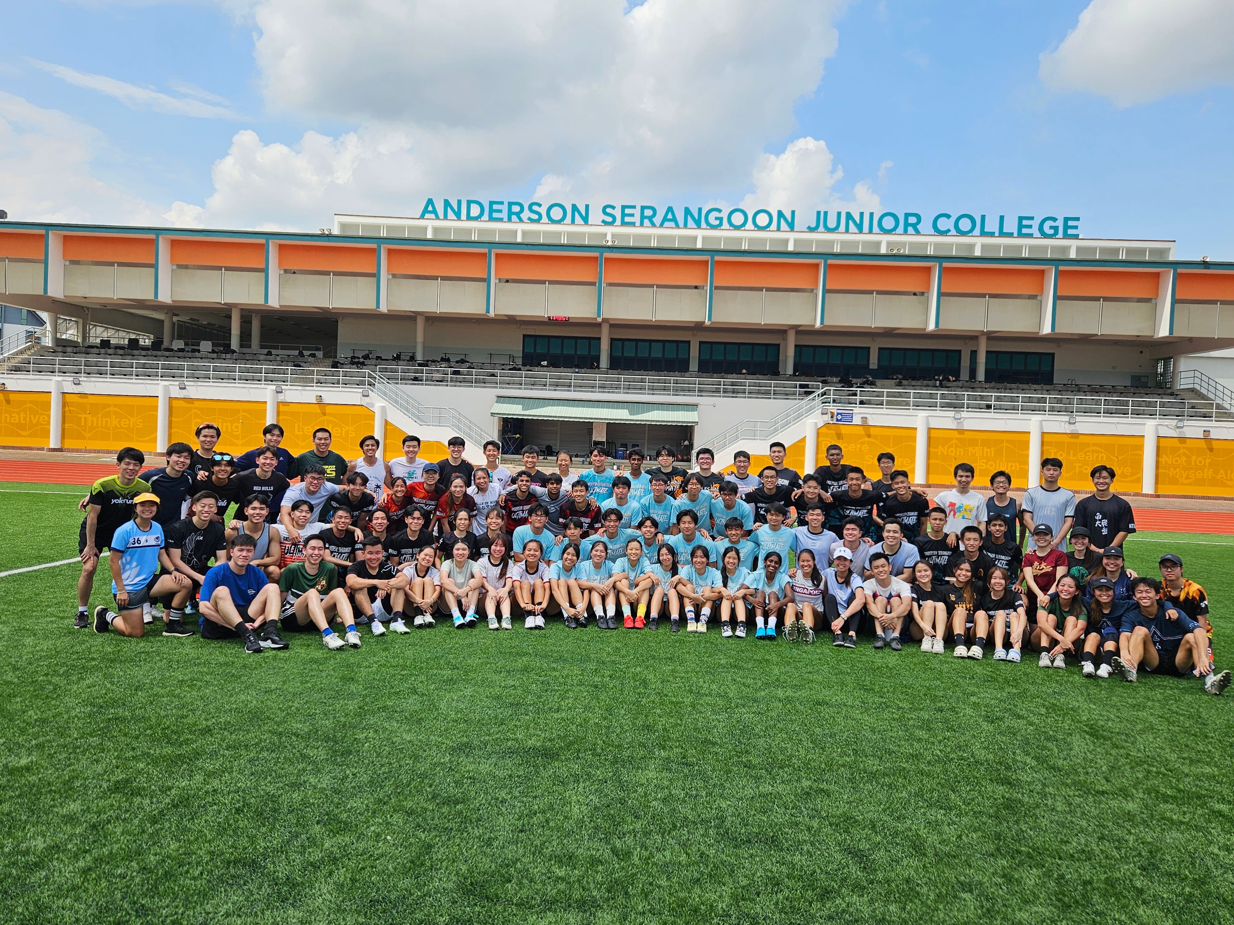 Large group seated on a sports field in front of a stadium labeled ANDERSON SERANGOON JUNIOR COLLEGE