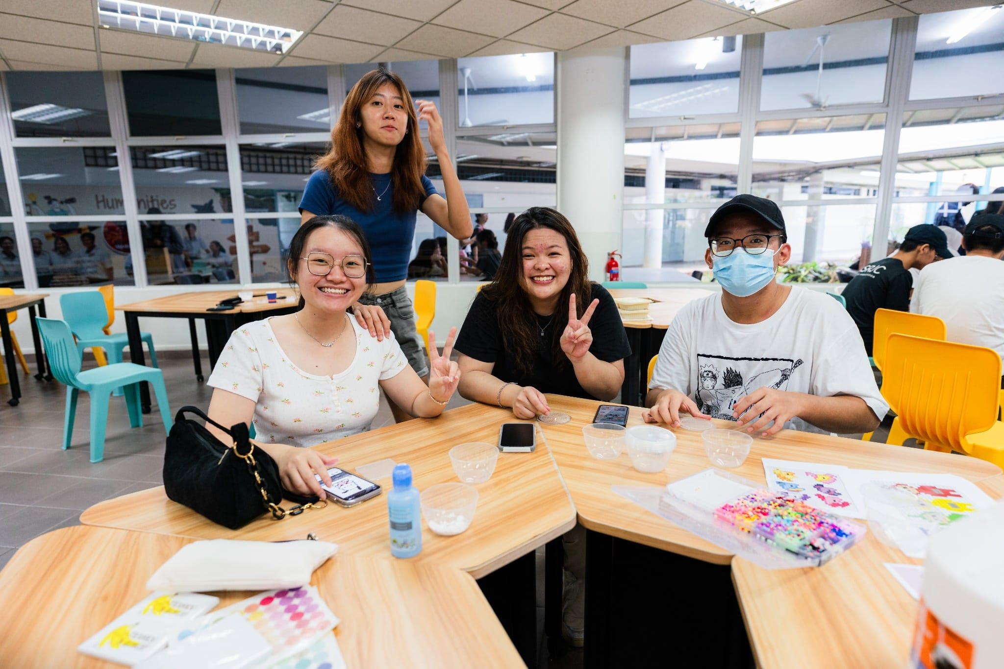 Four young people smile at a table with craft supplies and phones in a bright indoor space.