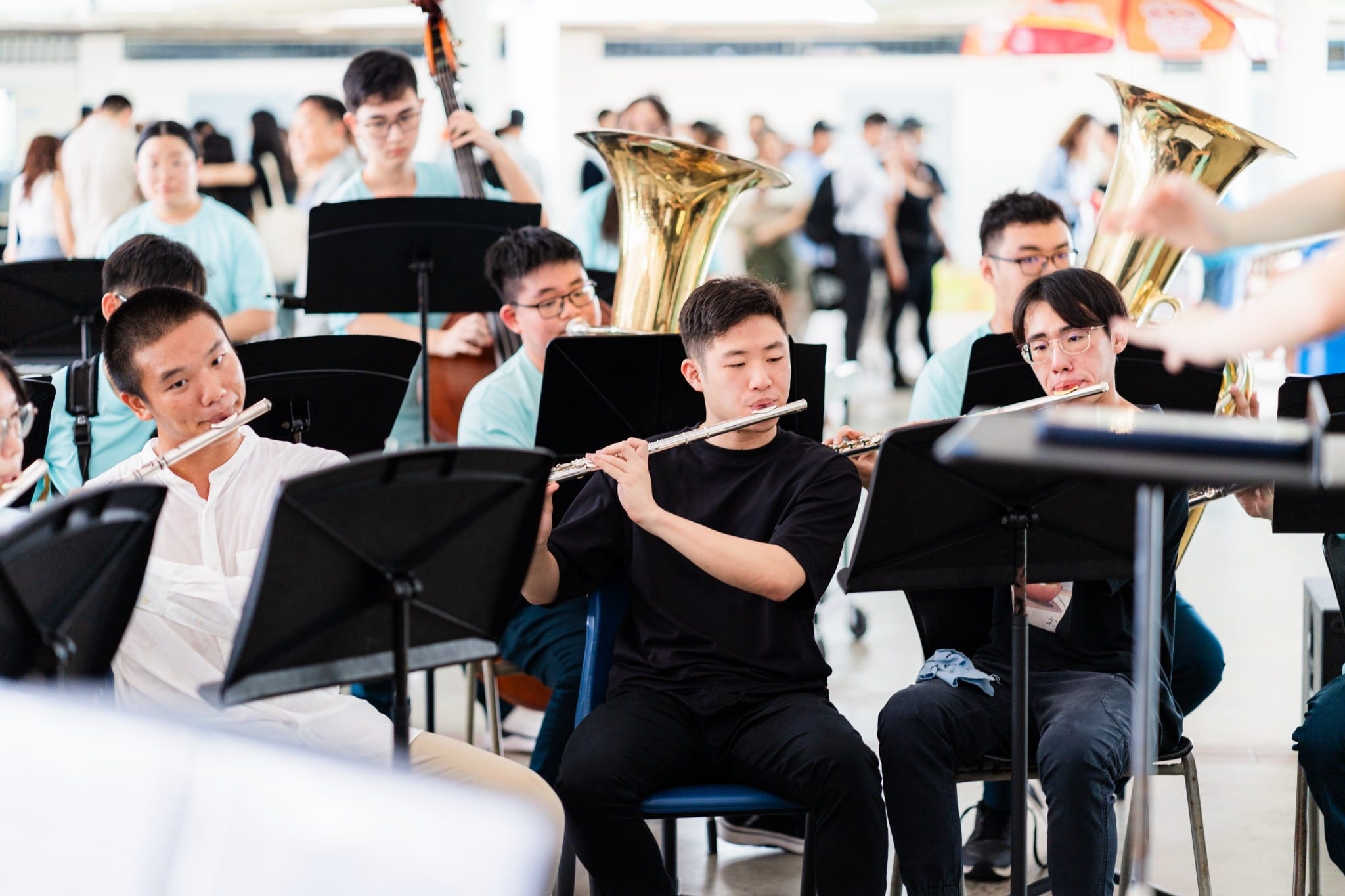 Young musicians seated with music stands play flutes, tubas, and a double bass during an indoor orchestra performance.