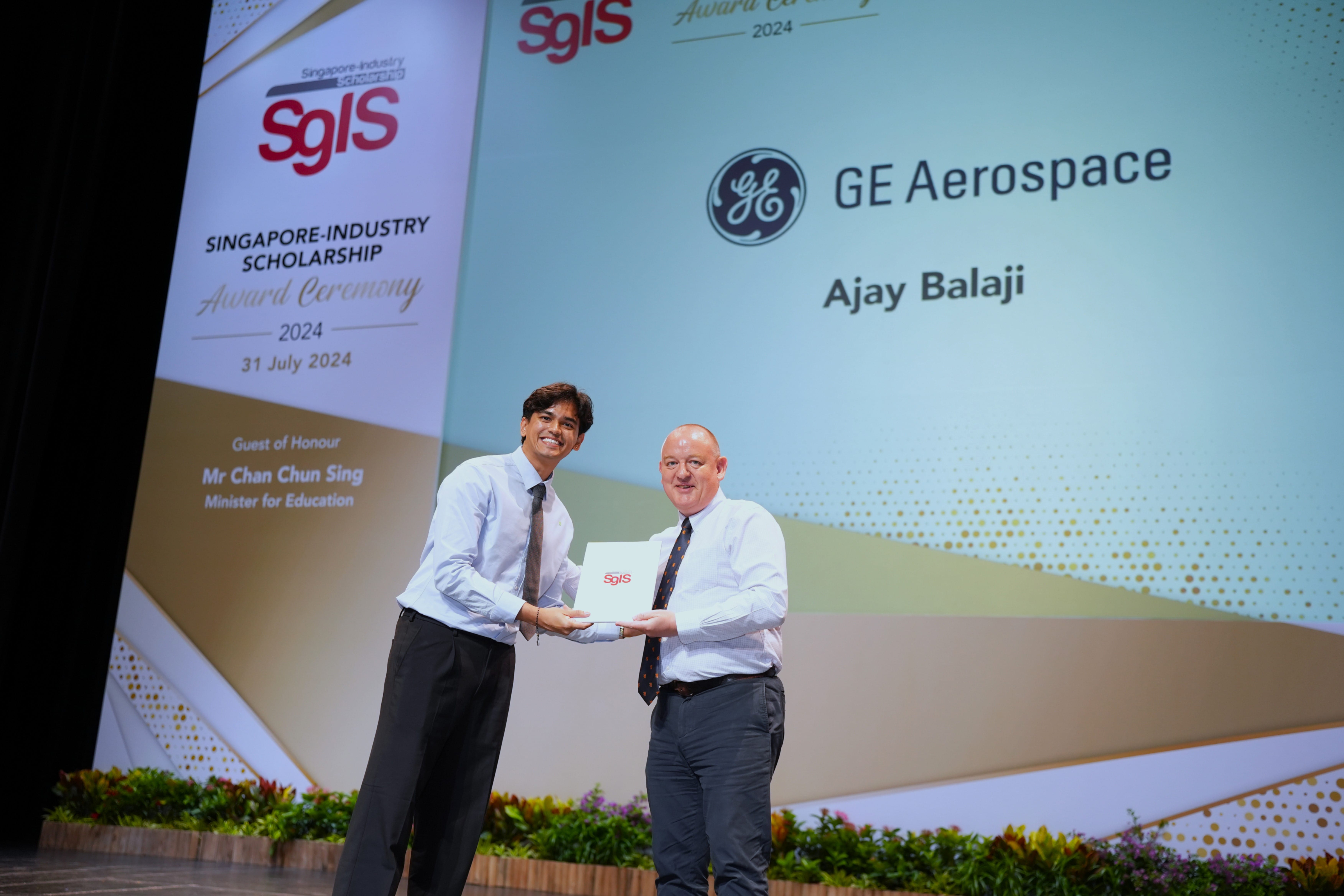 Two men on stage exchange a certificate at the 2024 Singapore-Industry Scholarship ceremony for Ajay Balaji from GE Aerospace.