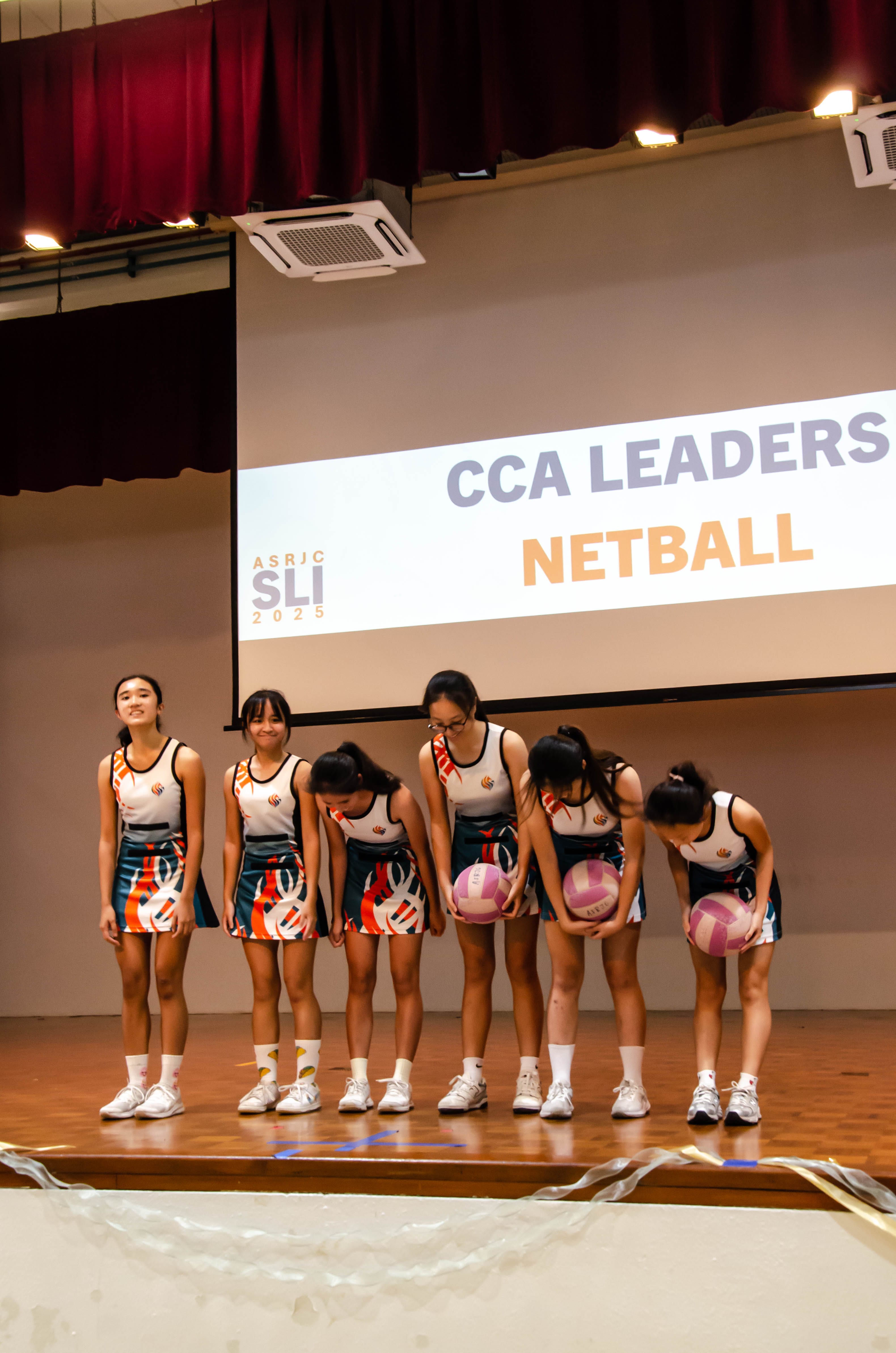 Six girls in netball uniforms bow on stage before a screen reading CCA Leaders Netball ASRJC SLI 2025.