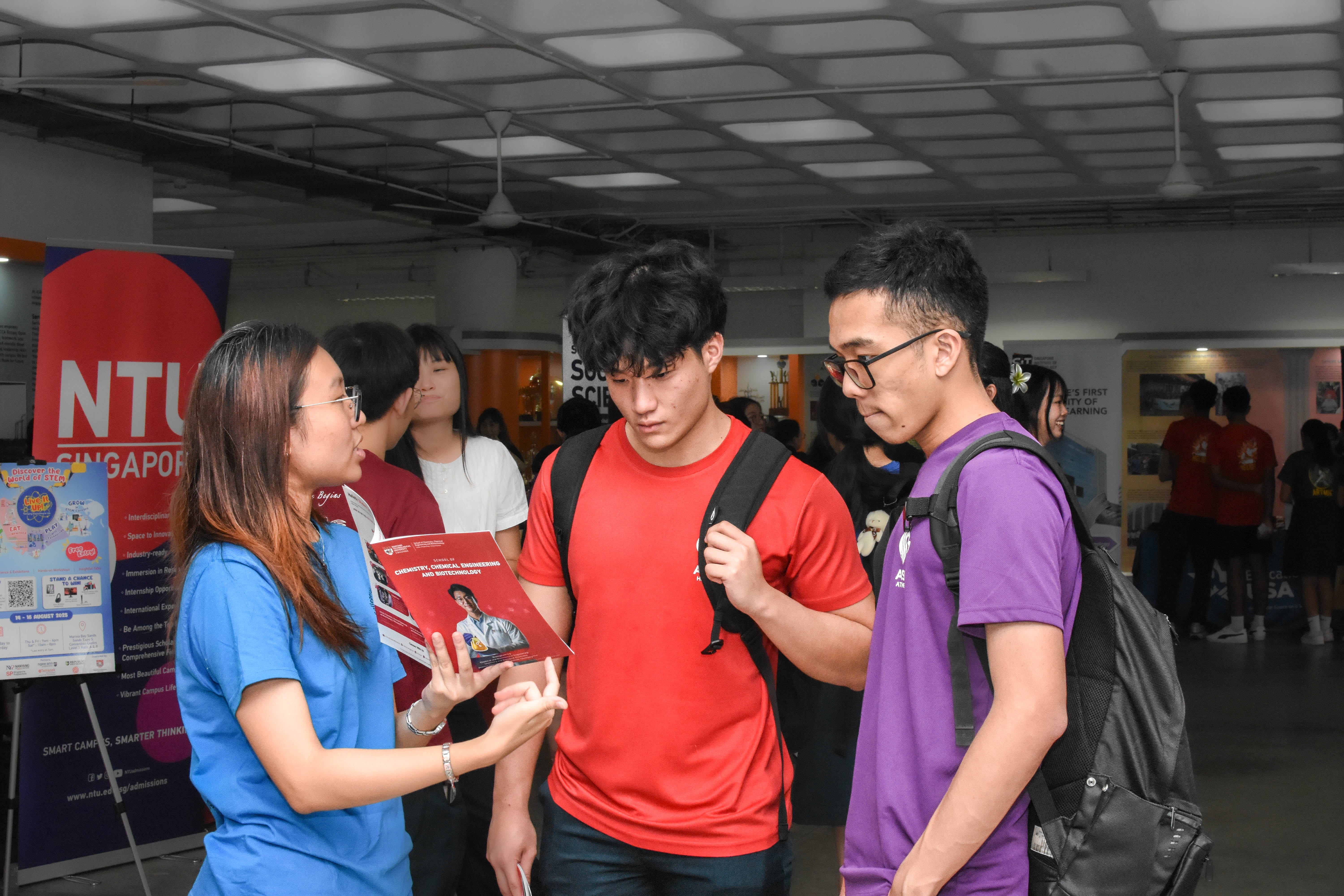 A woman holds an NTU brochure while talking to two male students at a university open day event.