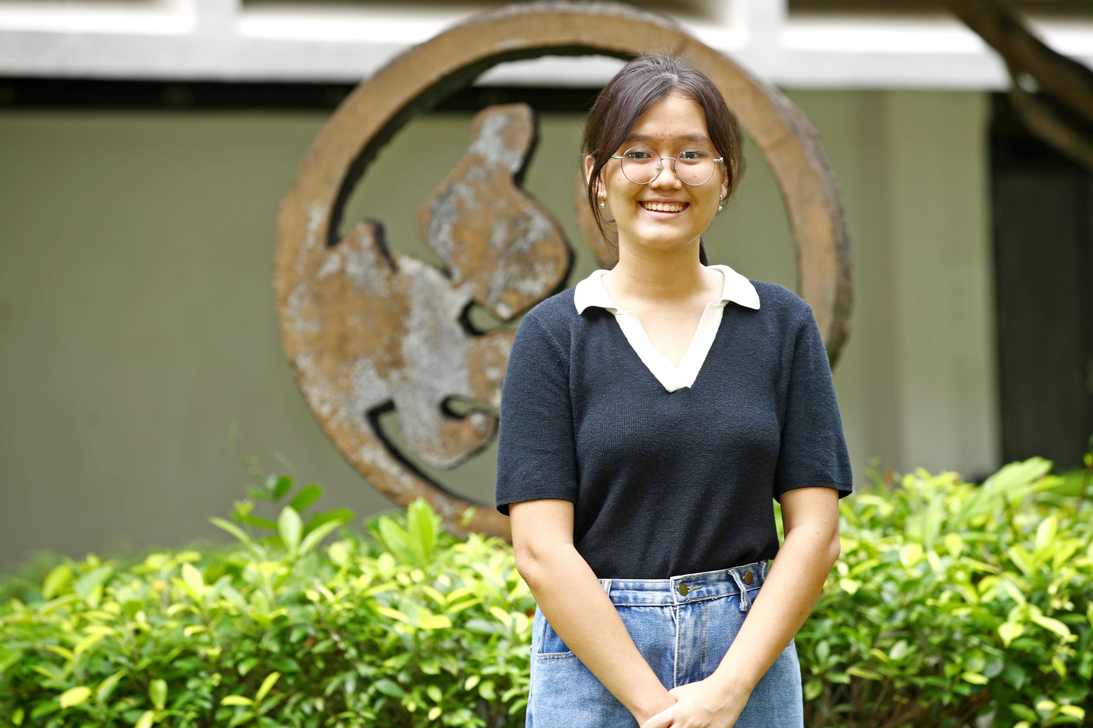 Smiling woman with glasses stands by green shrubs, with a large rusted circular sculpture behind her.