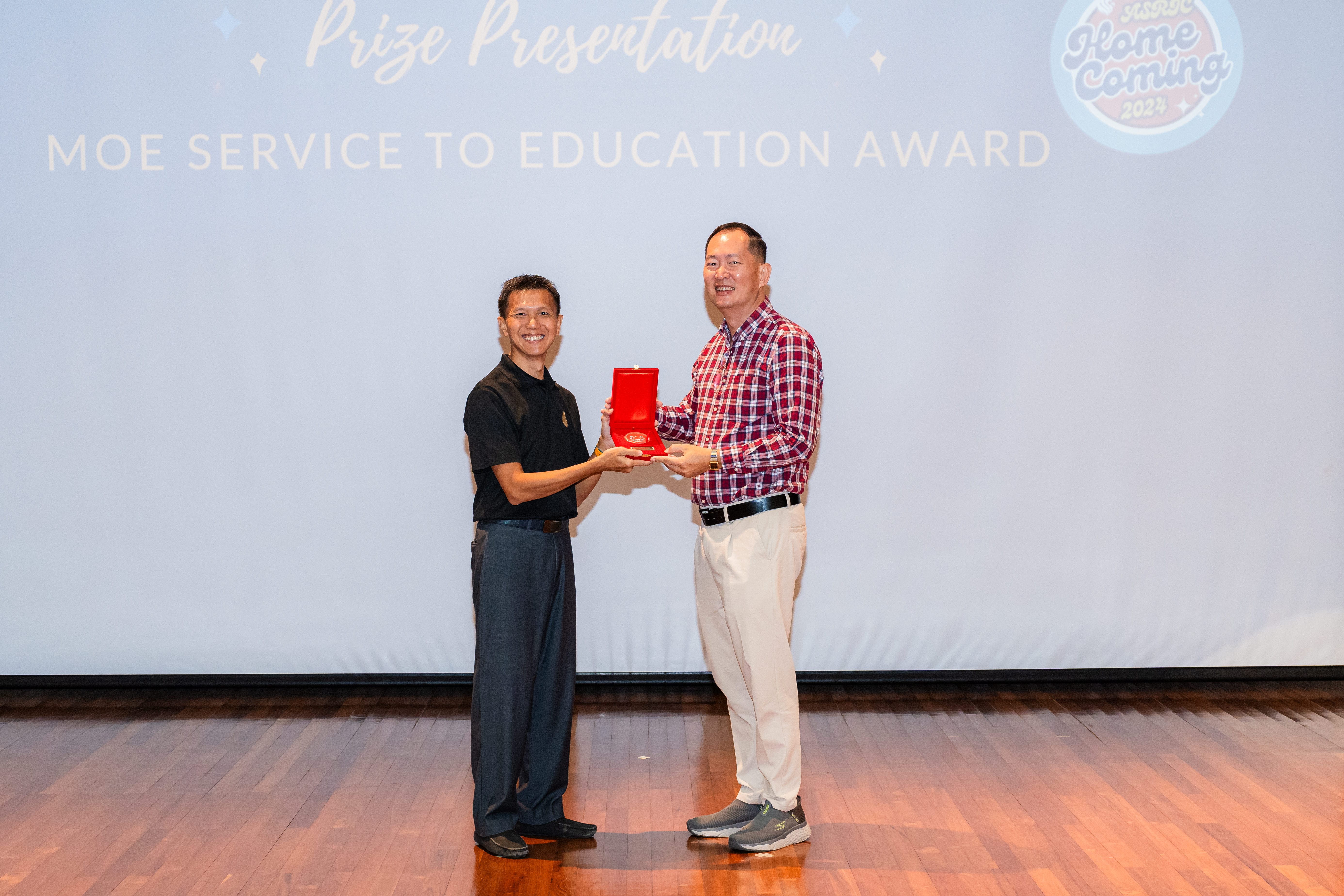 Two men hold a red award box on stage in front of a screen displaying MOE Service to Education Award.