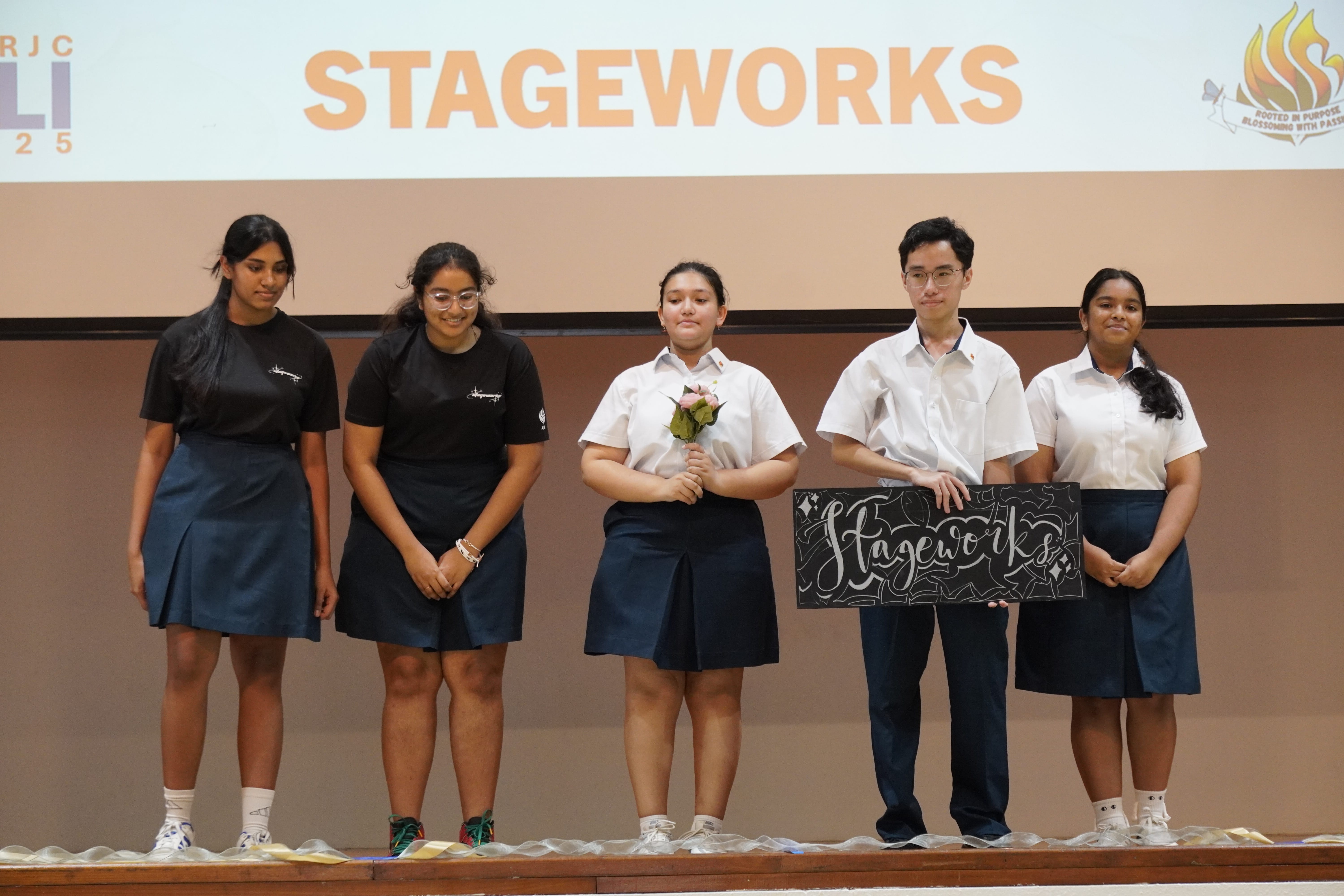 Five students on a stage beneath STAGEWORKS; one holds flowers and another holds a Stageworks sign.