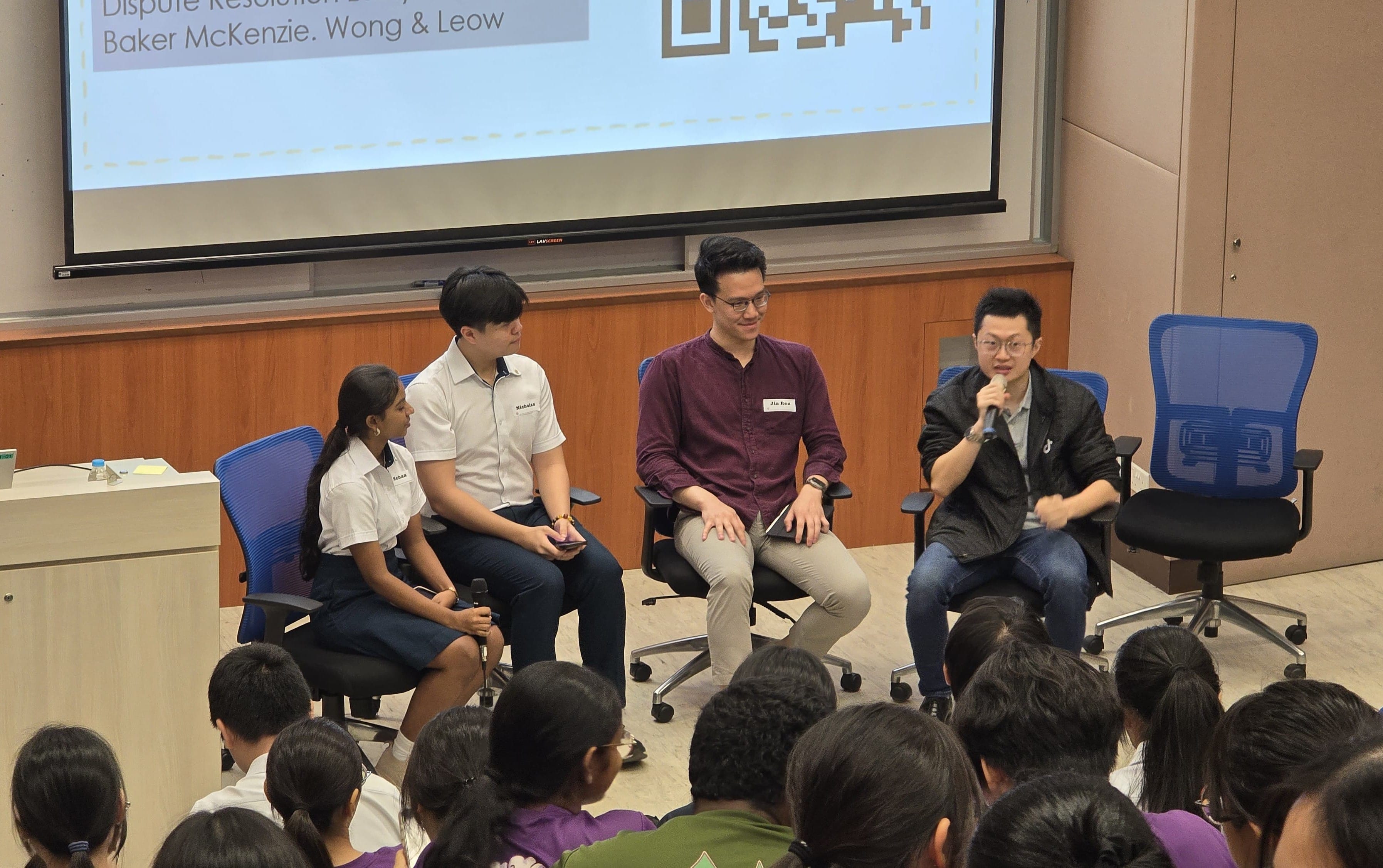 Four panelists sit on chairs facing an audience in a seminar room with a Baker McKenzie projection screen behind them.