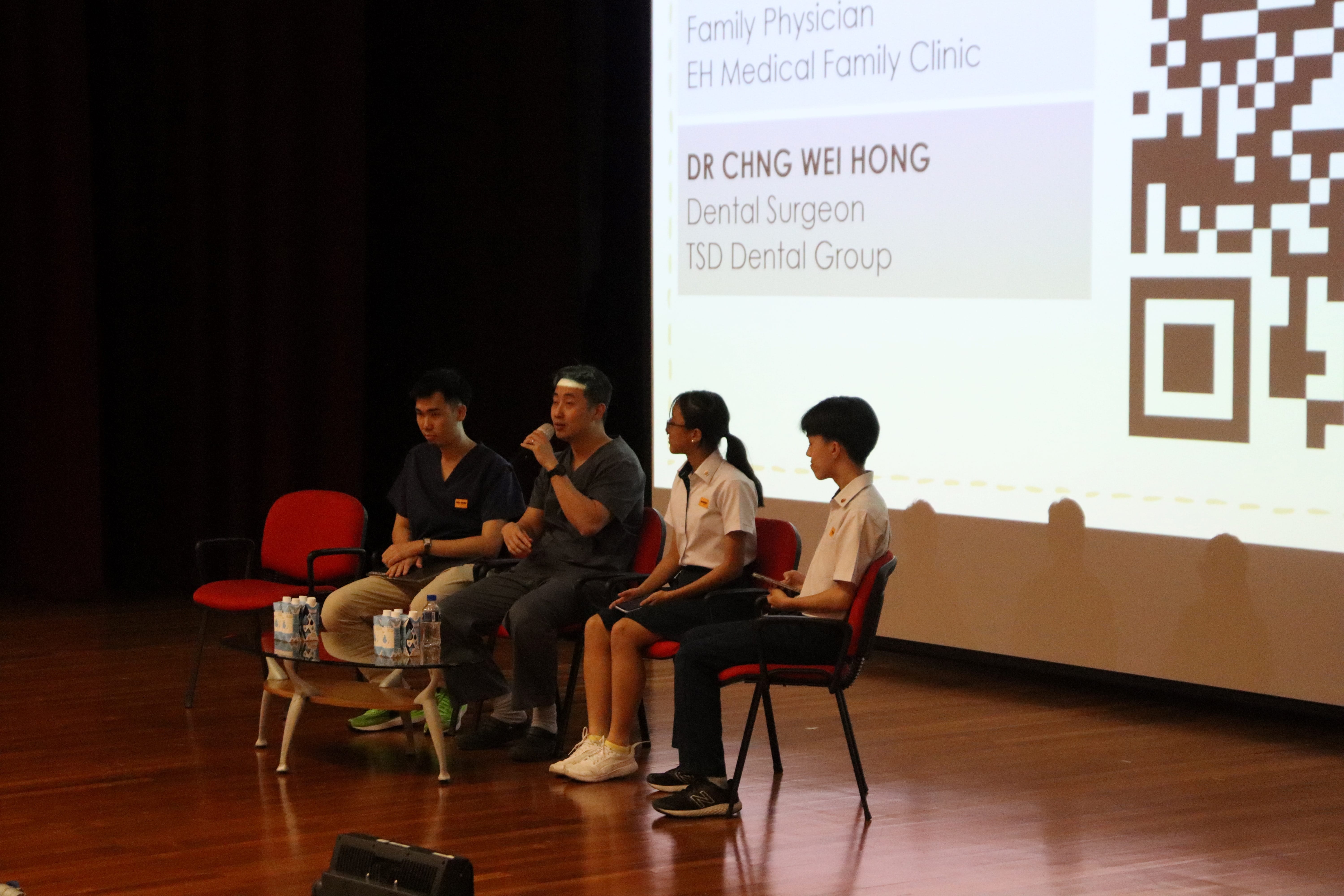 Four panelists seated on stage; one speaks into a mic beside a projected slide with a QR code.