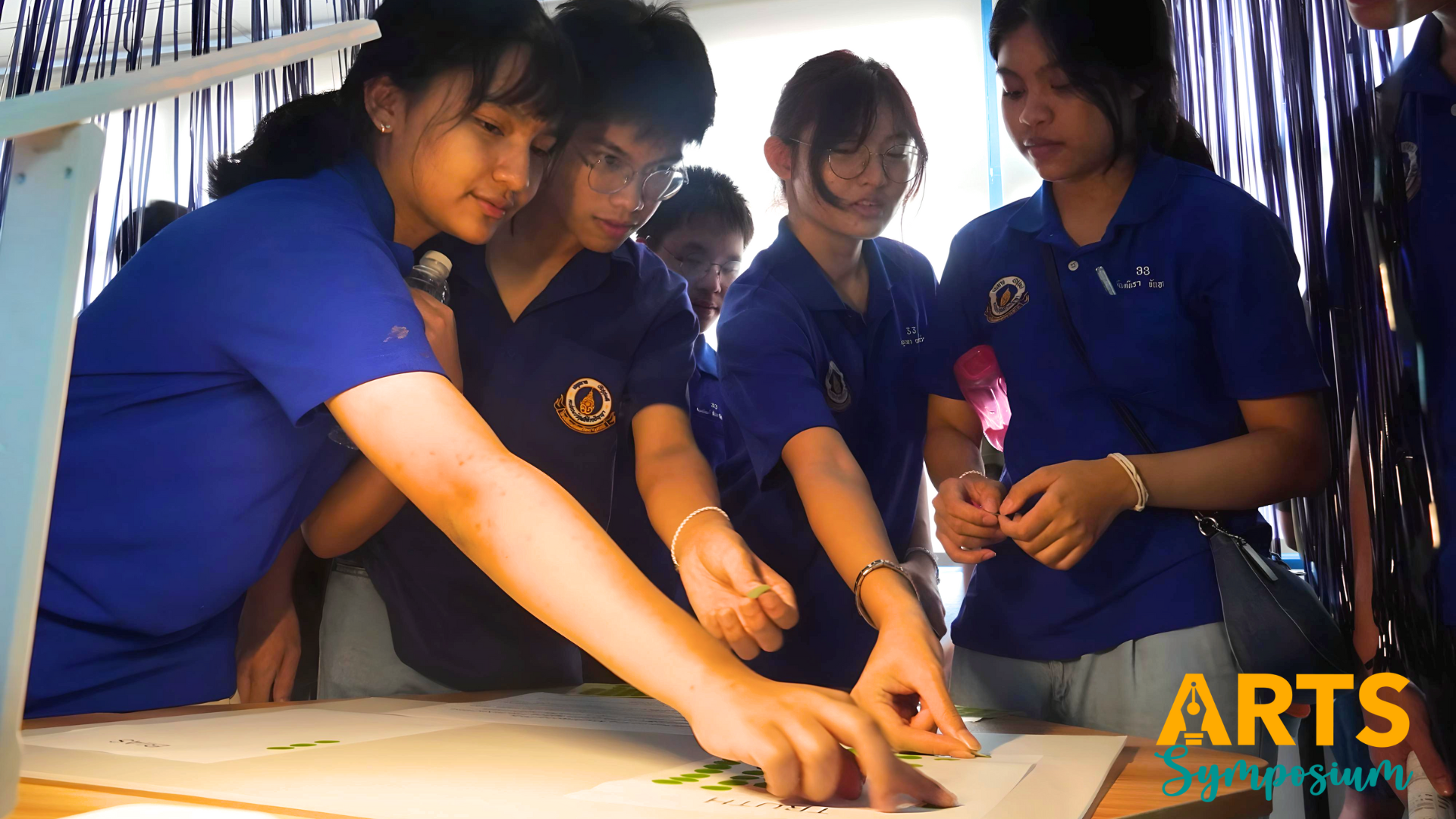 Several students in blue uniforms lean over a lit table, pointing at papers with small green designs.